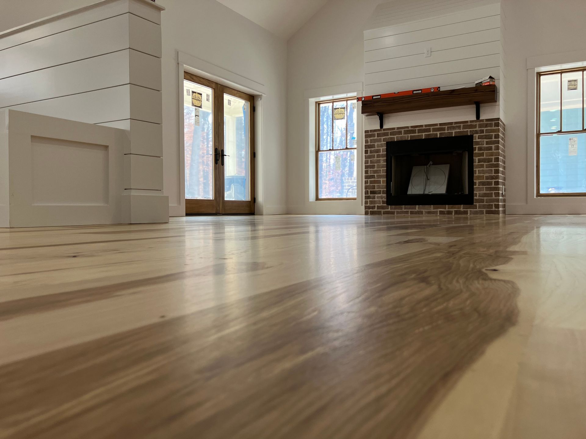 Empty living room with hardwood floors, white walls, large windows, and a brick fireplace