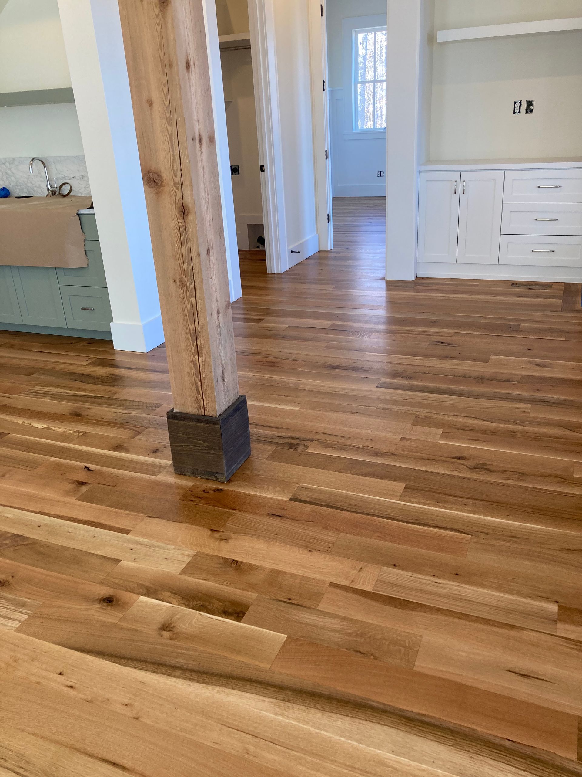 Empty hardwood living space with exposed wooden posts, white cabinetry, and a hallway in the background
