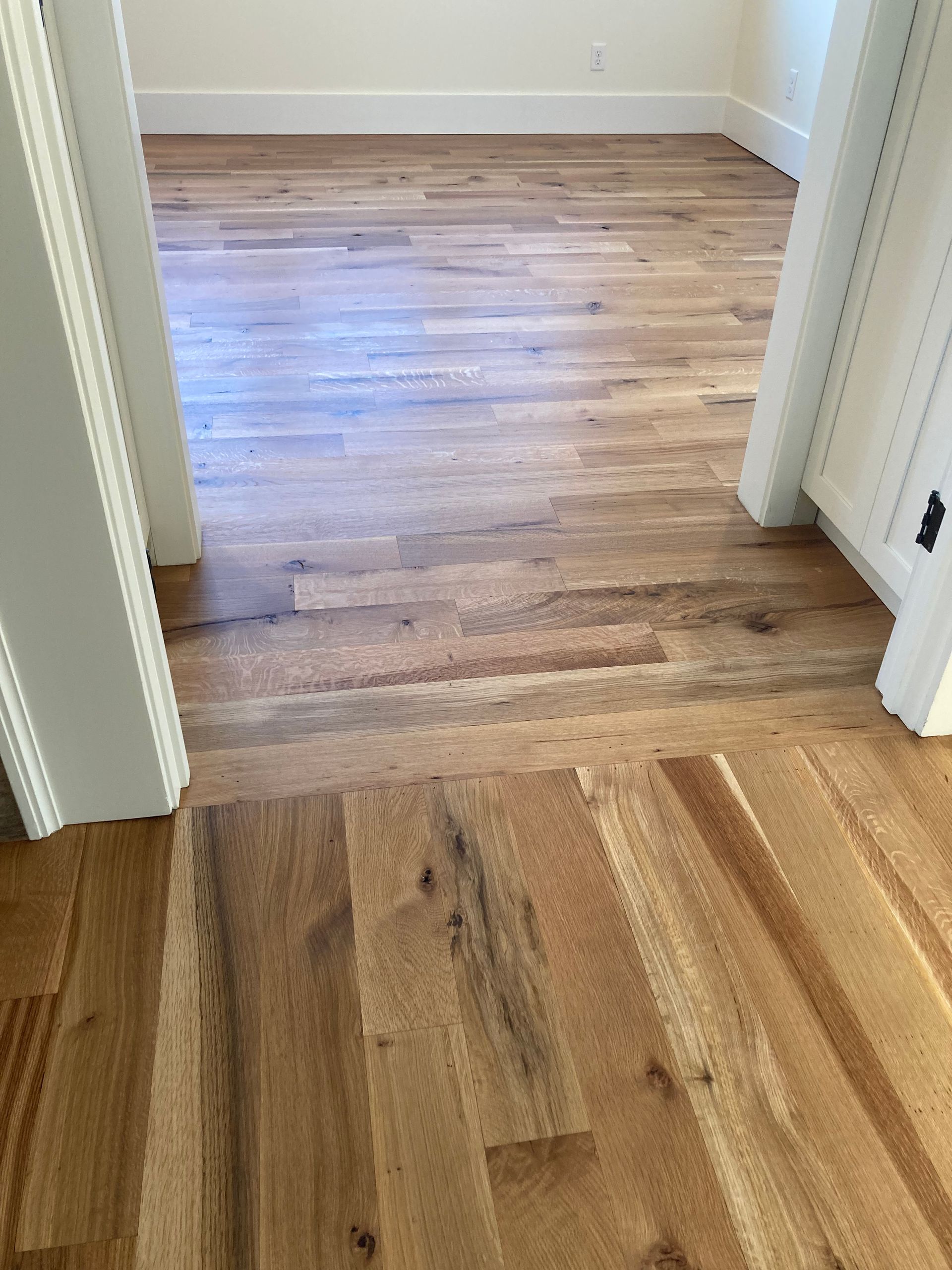 Wooden flooring with a transition seam between a hallway and a room, viewed through a doorway.