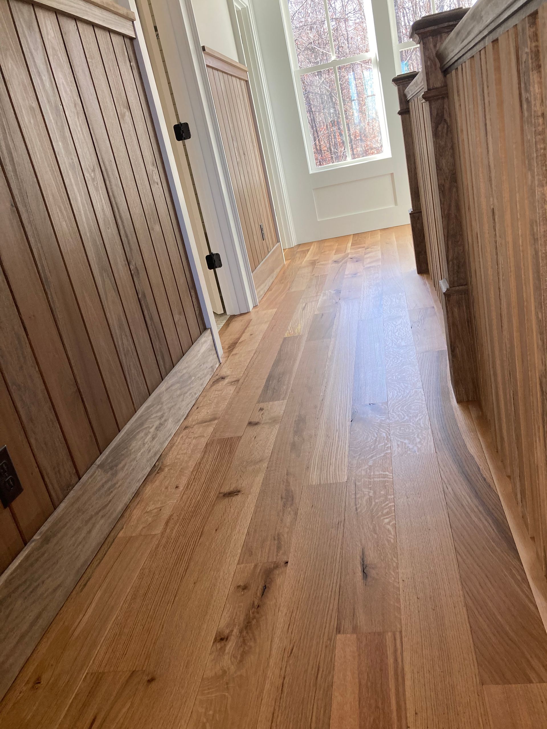 Narrow hallway with wood floors, paneled walls, and a bright door at the end.