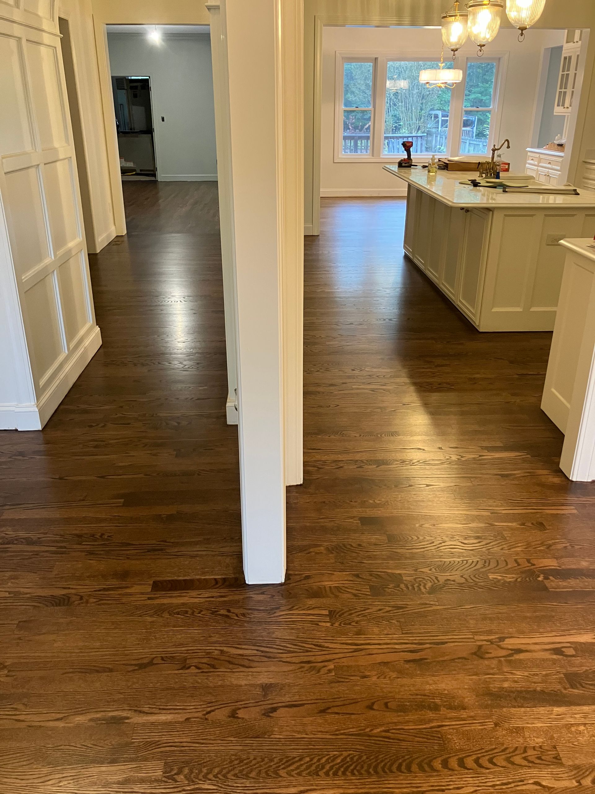 Empty hallway and kitchen with dark hardwood floors, white walls, and large windows with daylight.