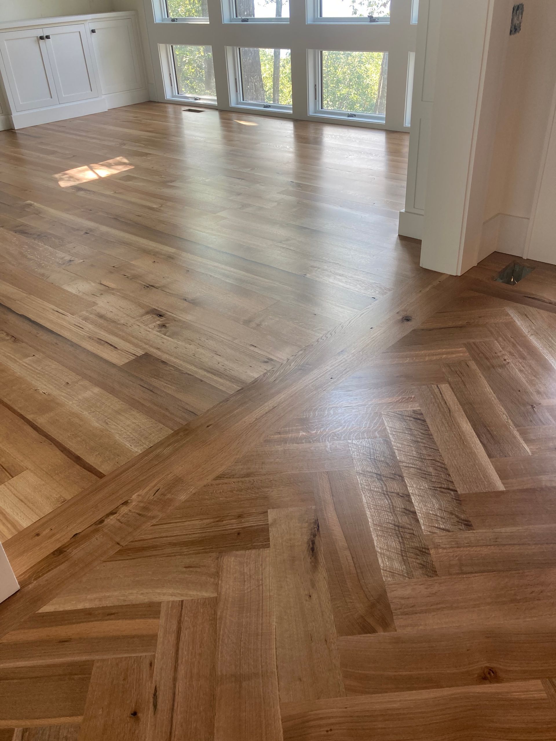 Sunlit empty room with light hardwood and herringbone wood floor near large windows