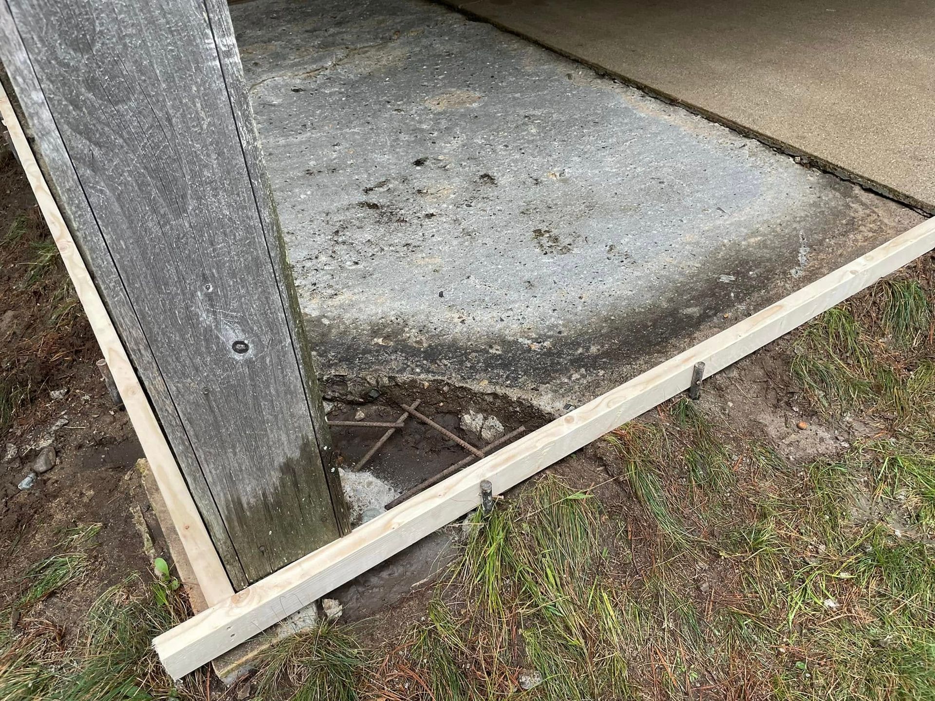 Concrete porch corner with a cracked base and a leaning wooden support post beside grass and dirt.