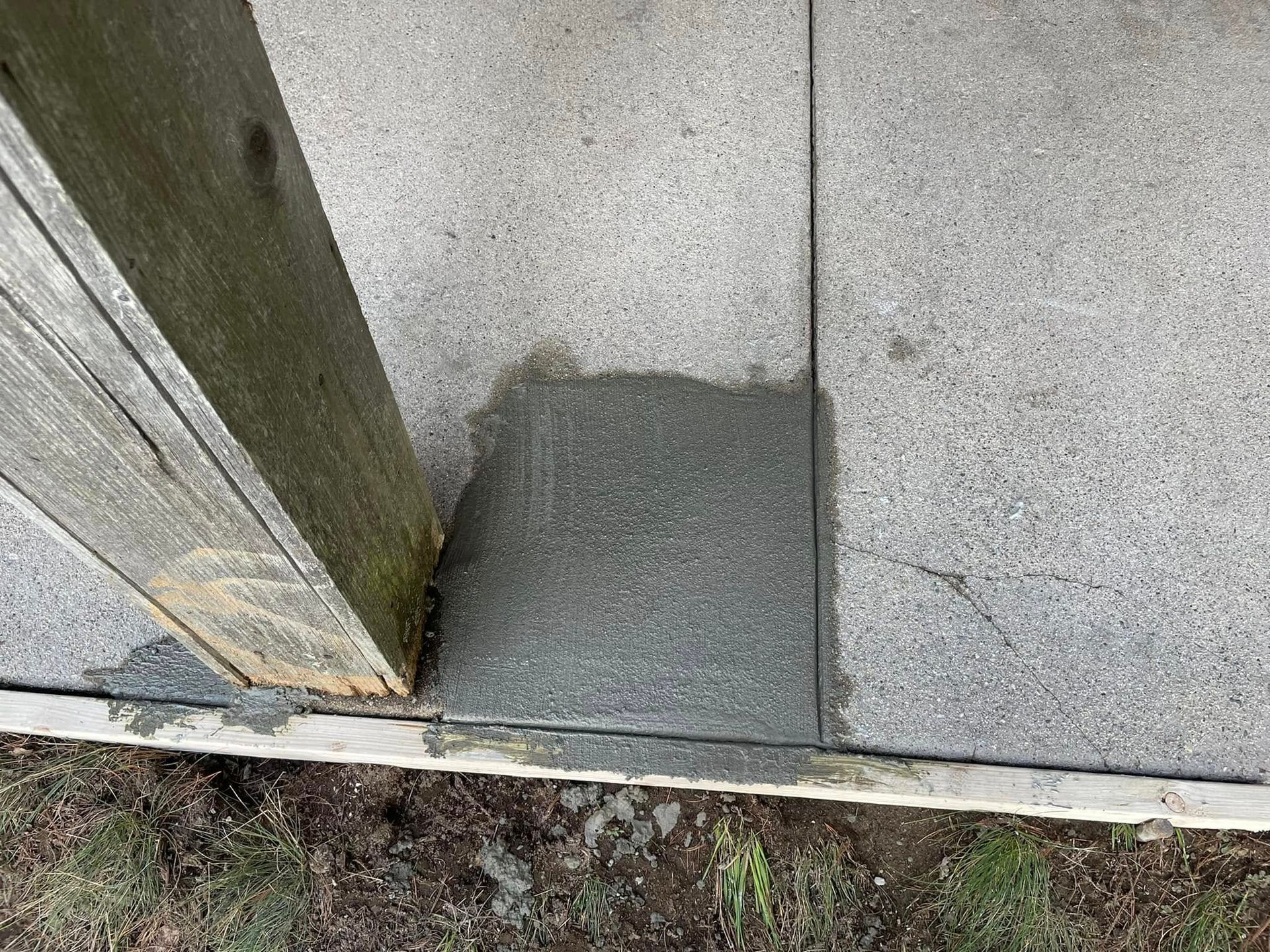 Concrete repair patch at the base of a wall beside a wooden post outdoors.