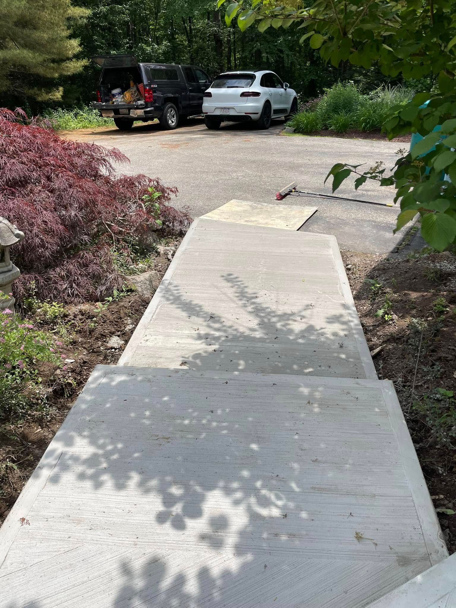 Concrete sidewalk leading to a driveway with two parked trucks and leafy bushes