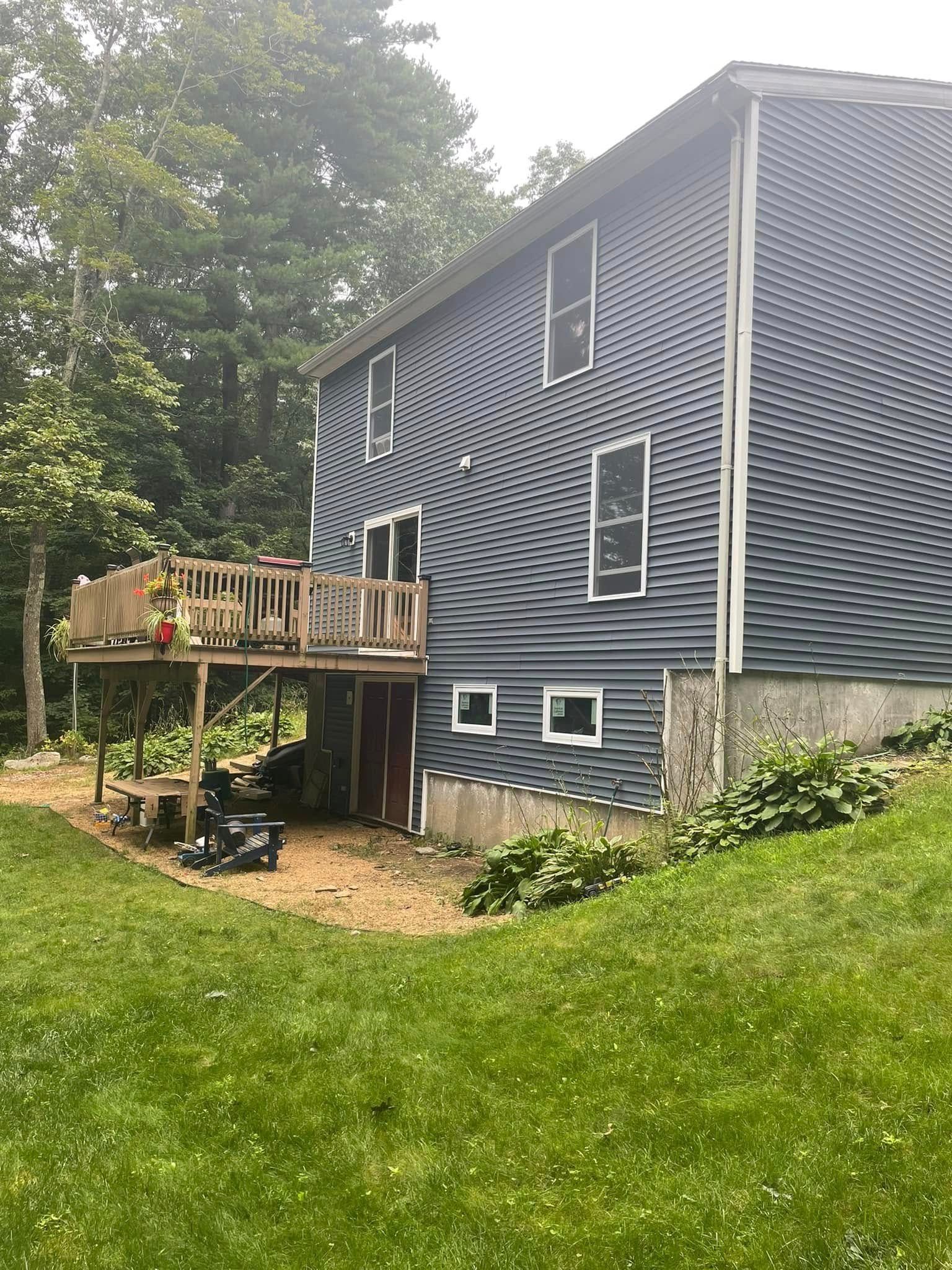 Two-story dark house with a wooden deck, set on a grassy slope beside trees.