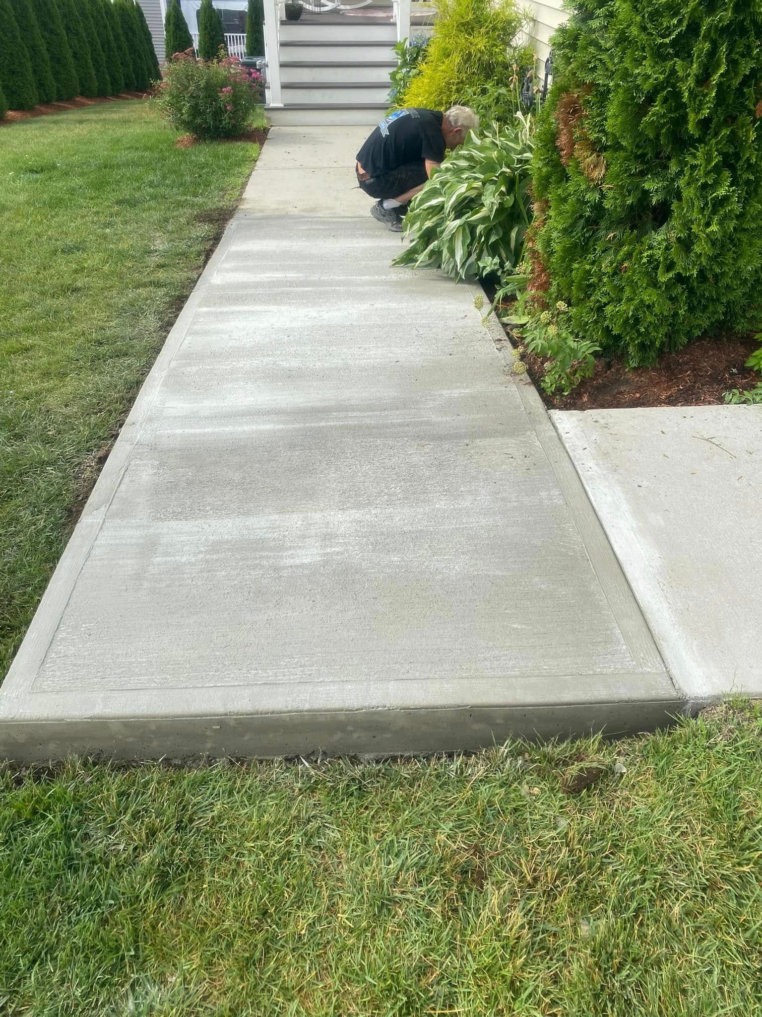 Person kneeling beside a concrete walkway, trimming bushes in a front yard.