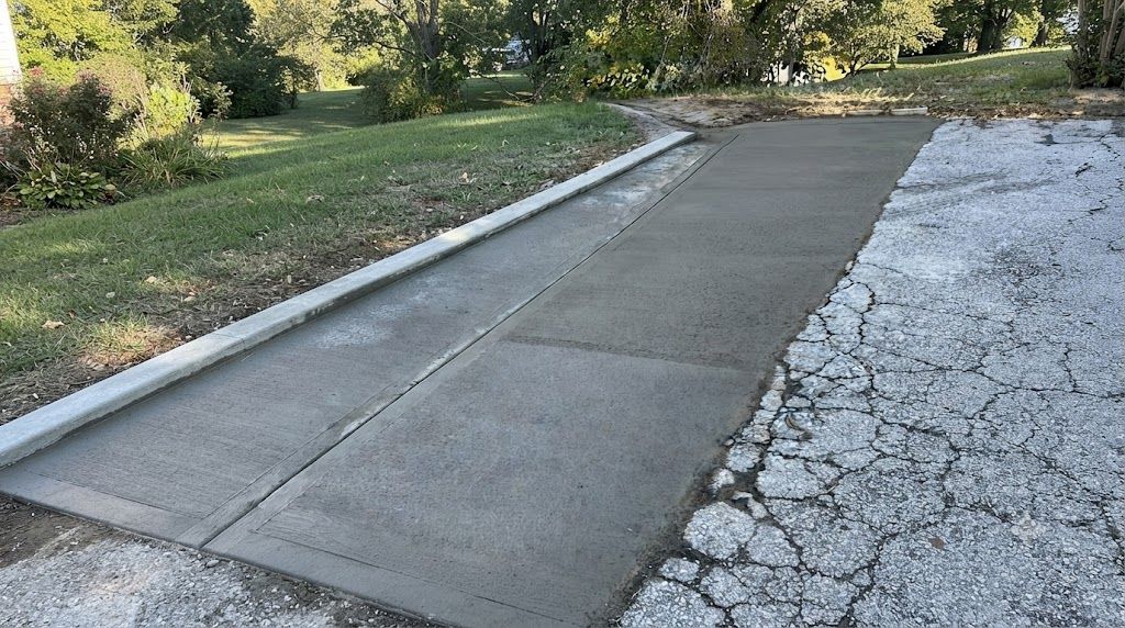 Freshly poured concrete driveway beside a cracked, weathered path with grass and trees nearby