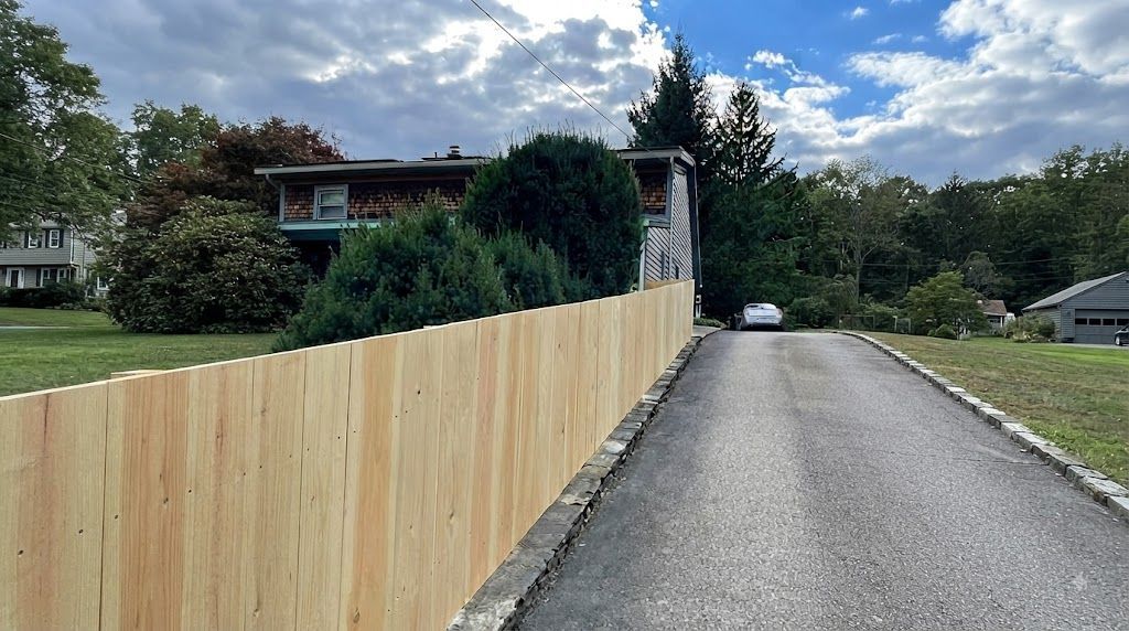 Suburban road beside a long wooden fence, with a house and trees under cloudy skies.