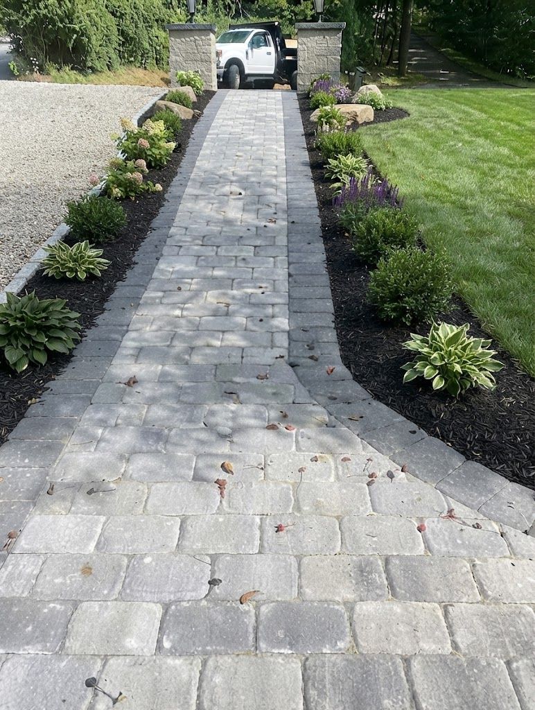 Stone driveway path between landscaped flower beds leading to a parked white truck and stone gate pillars