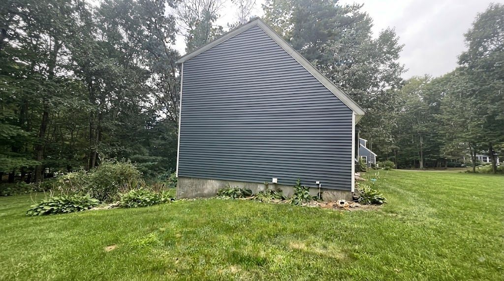 Gray house side with wavy siding, small flower bed, and green lawn bordered by trees.