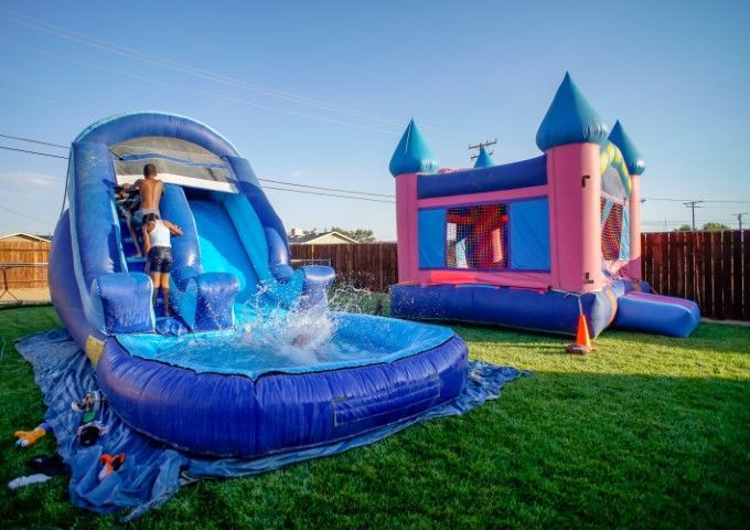 Blue and pink inflatable bounce house with a water slide on grass, with a child playing inside