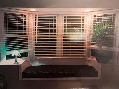 Cozy bay window seat with white blinds, dark cushion, side tables, and a potted plant at night