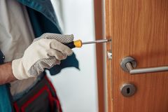 Gloved hand using a screwdriver to remove a door lock on a wooden door