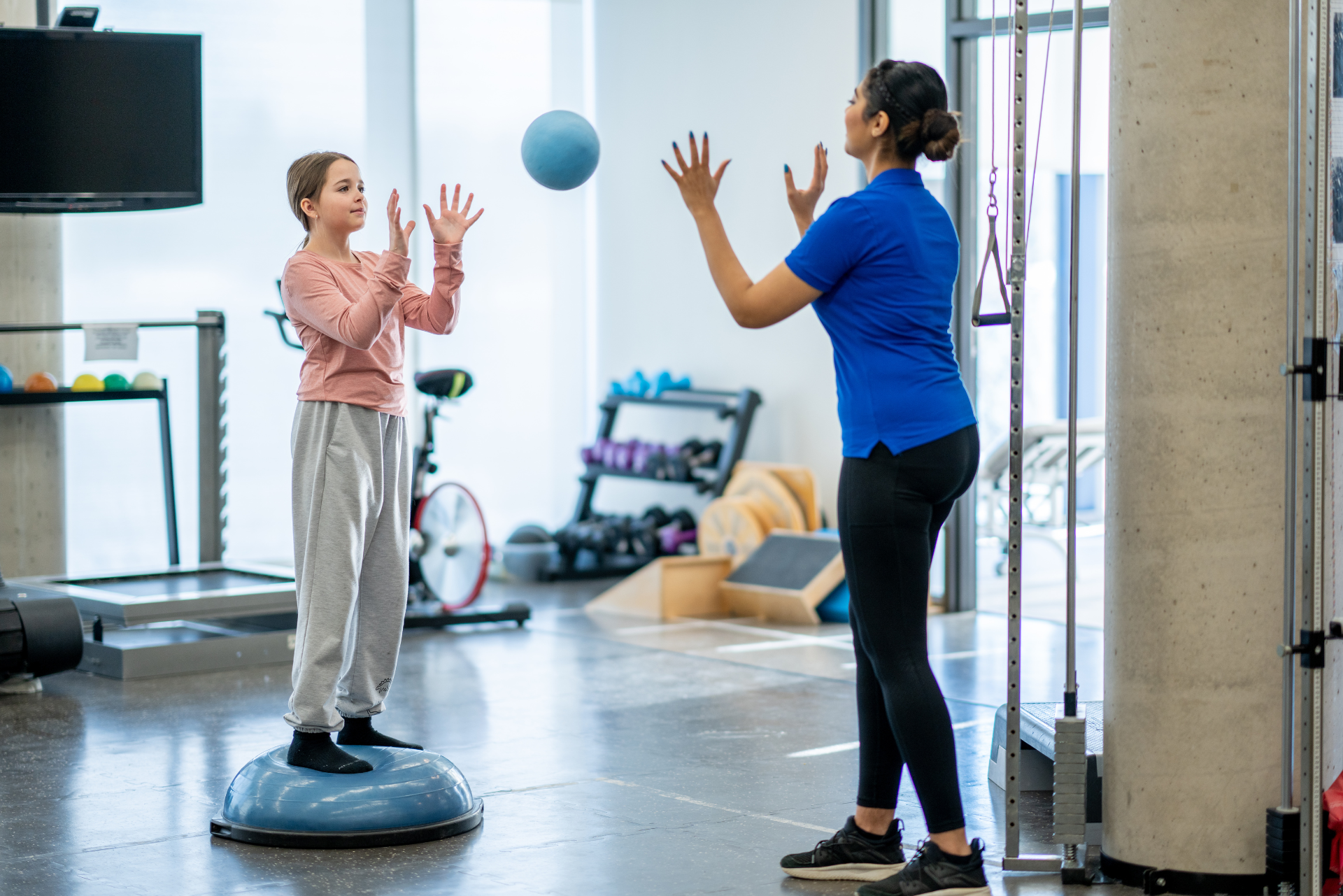 Une fille est en équilibre sur un ballon Bosu bleu et lance le ballon à une femme dans une salle de sport. La femme est vêtue de bleu, la fille de rose.