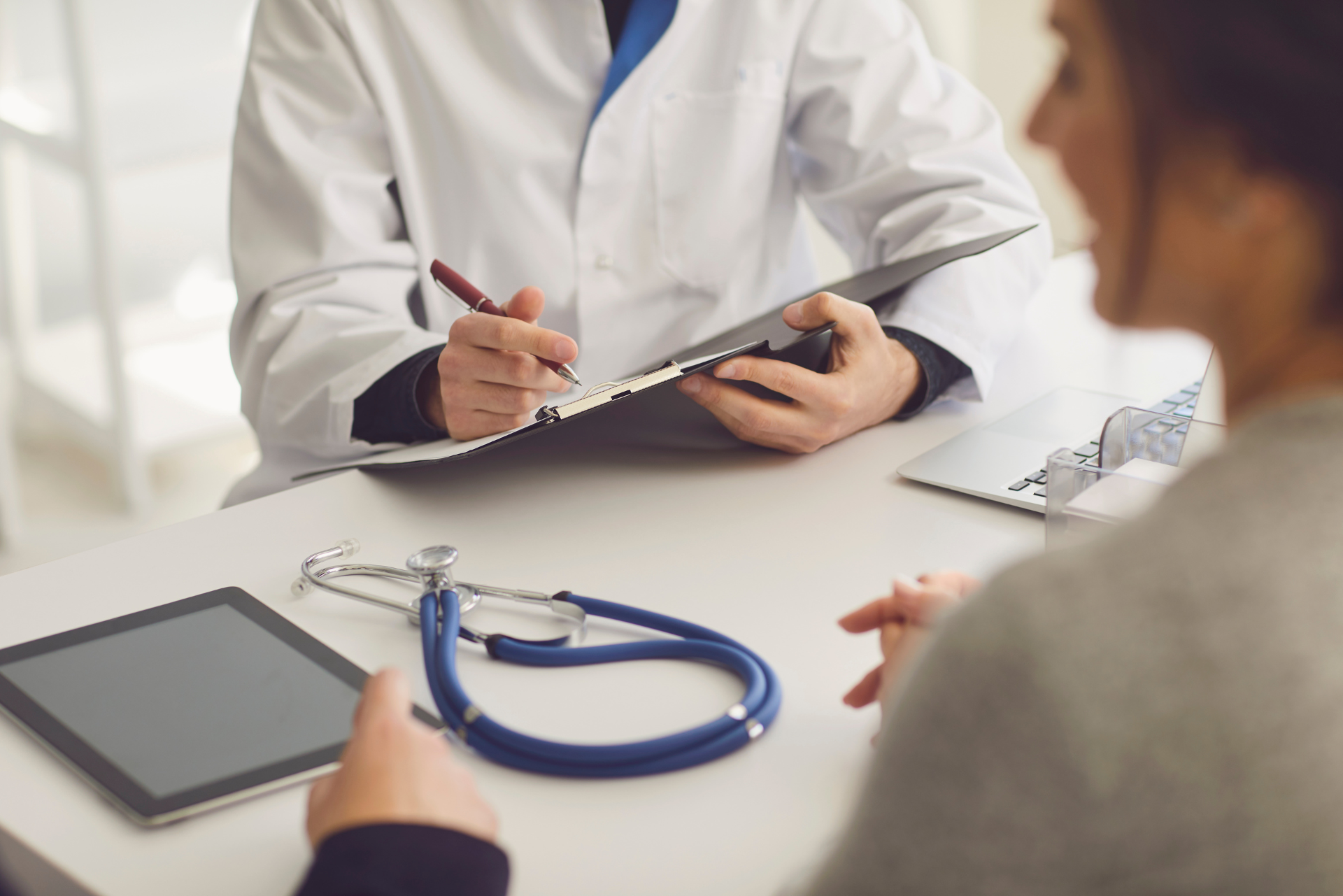 Médecin en blouse blanche écrivant sur un presse-papiers, parlant avec un patient à un bureau avec un stéthoscope et une tablette.