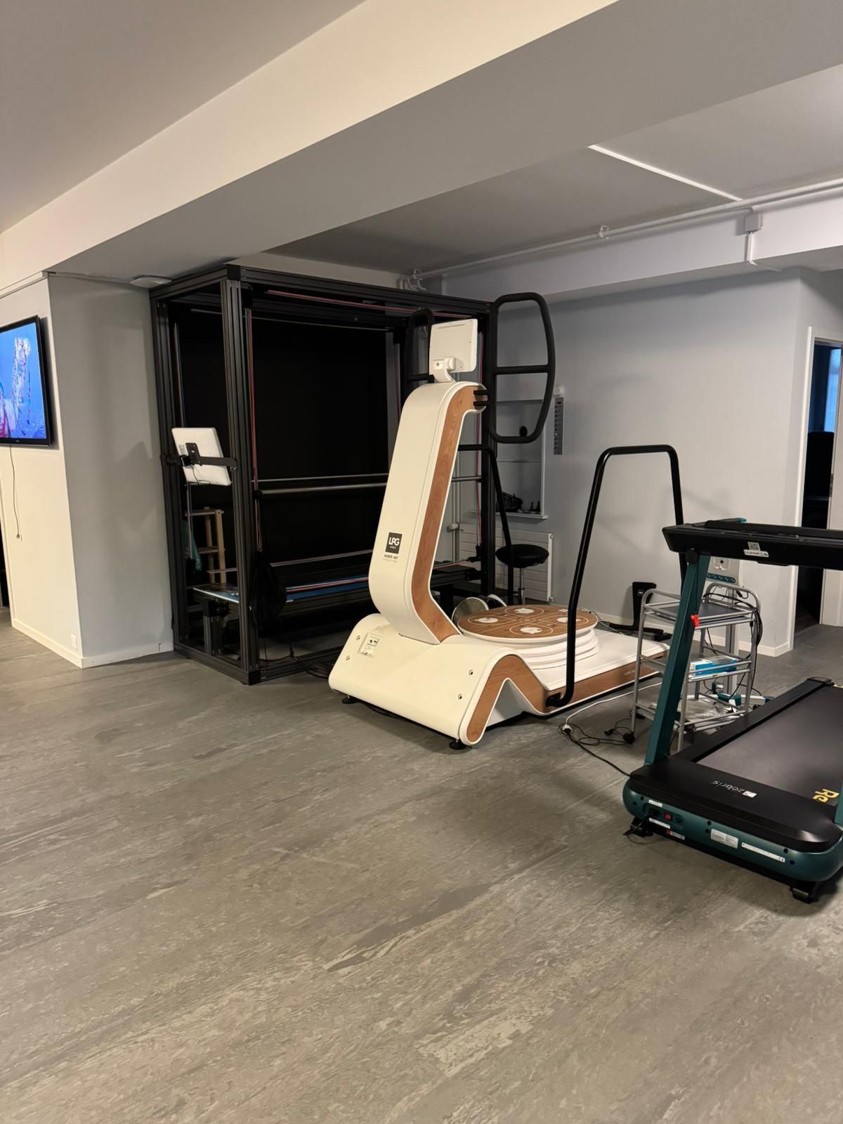 Une salle de sport avec des équipements : une machine blanche et marron, un tapis de course et une structure noire. Le sol est gris et les murs blancs.