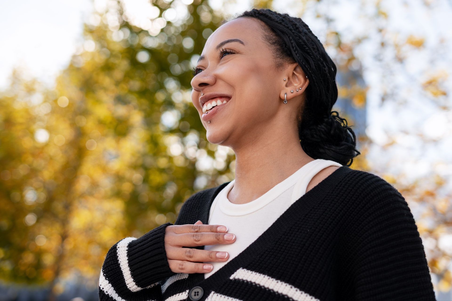 Smiling person outdoors in a black sweater, hand on chest, with autumn trees in the background