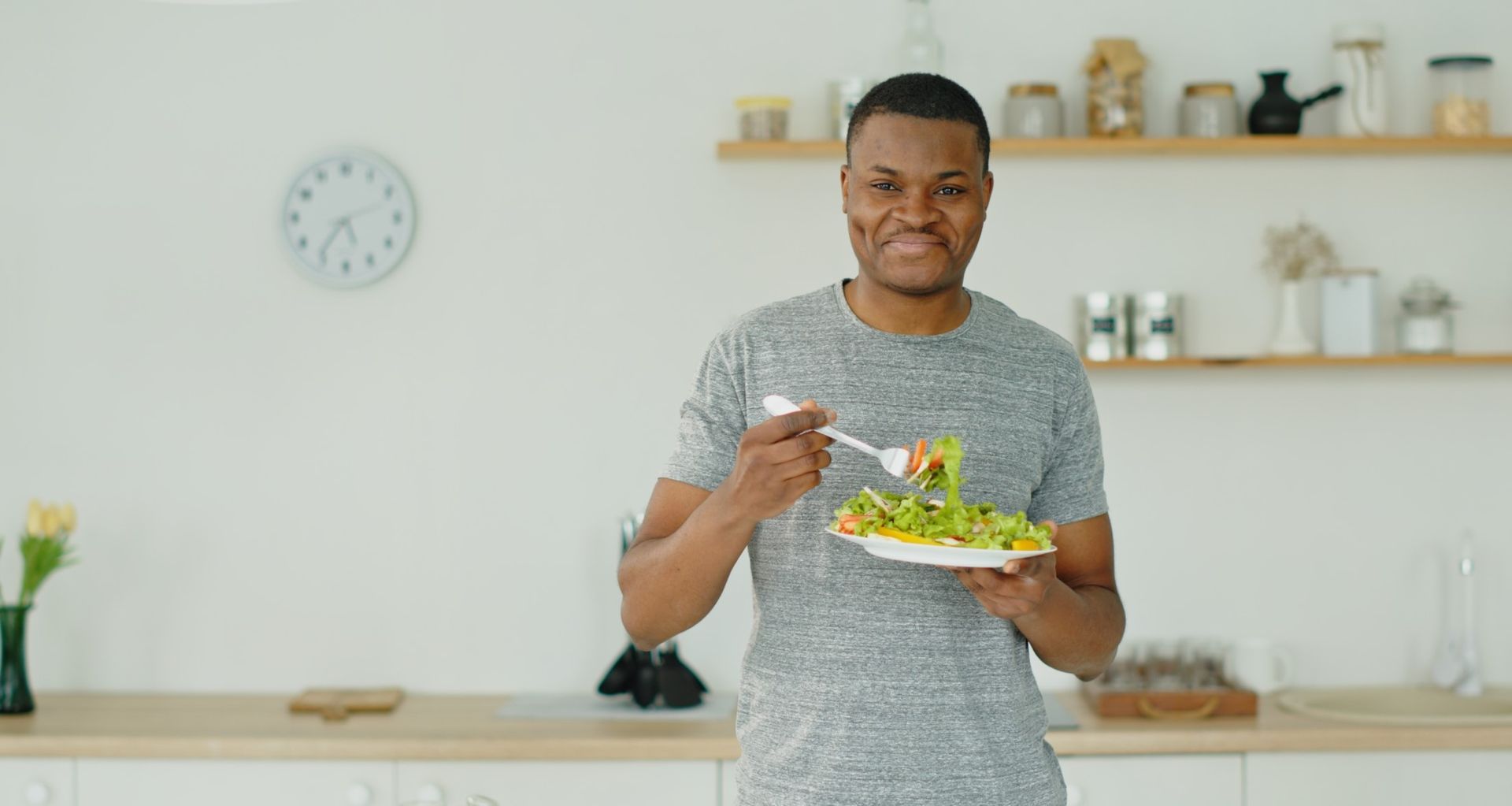Person holding a plate and spoon in a bright kitchen, with a clock and shelves in the background