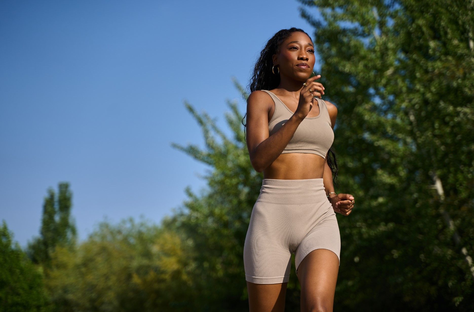 Woman jogging outdoors in a beige athletic outfit on a sunny day, with trees and blue sky behind her