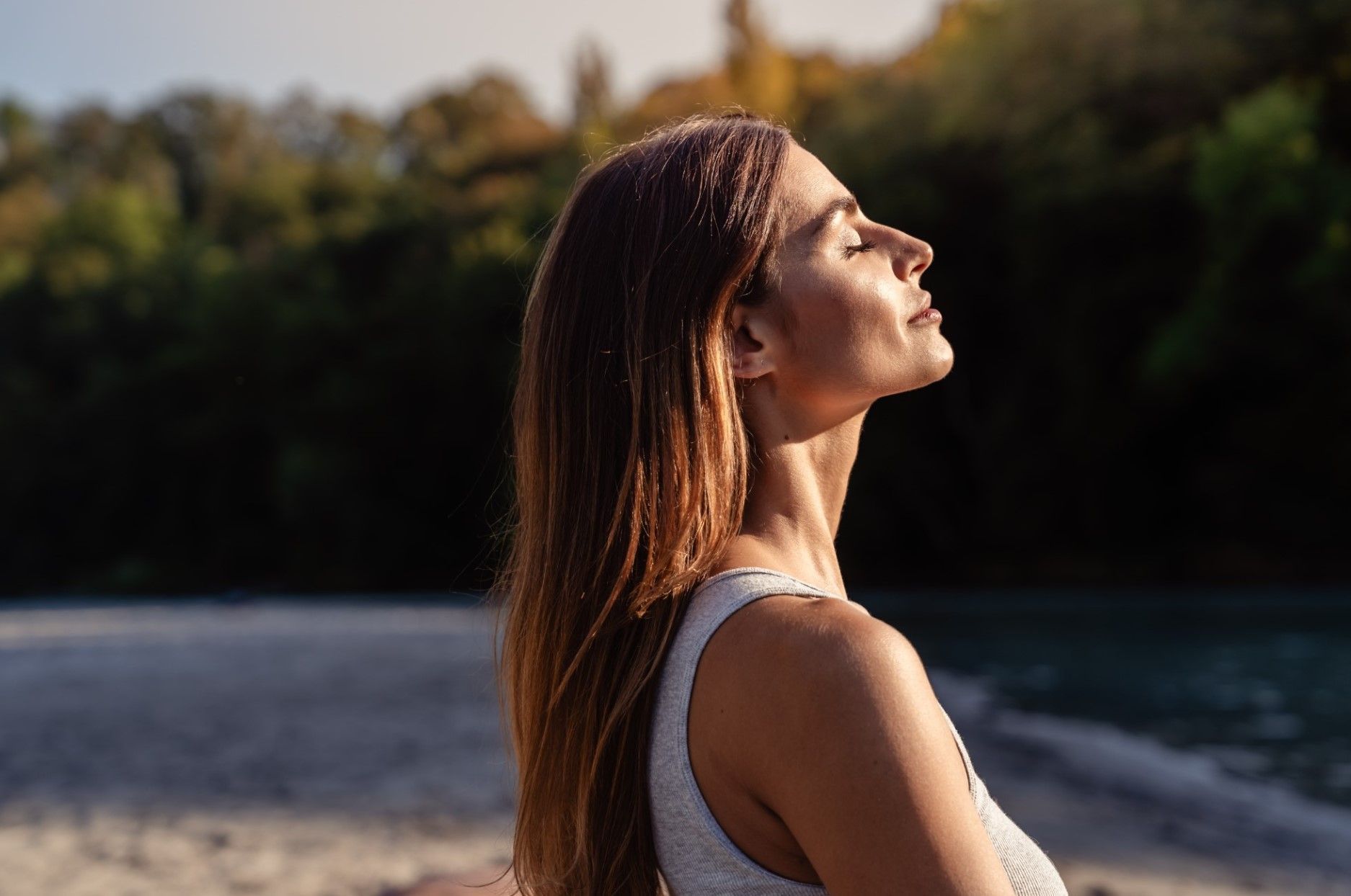 Woman in a white tank top standing by a river, looking up in warm sunlight.
