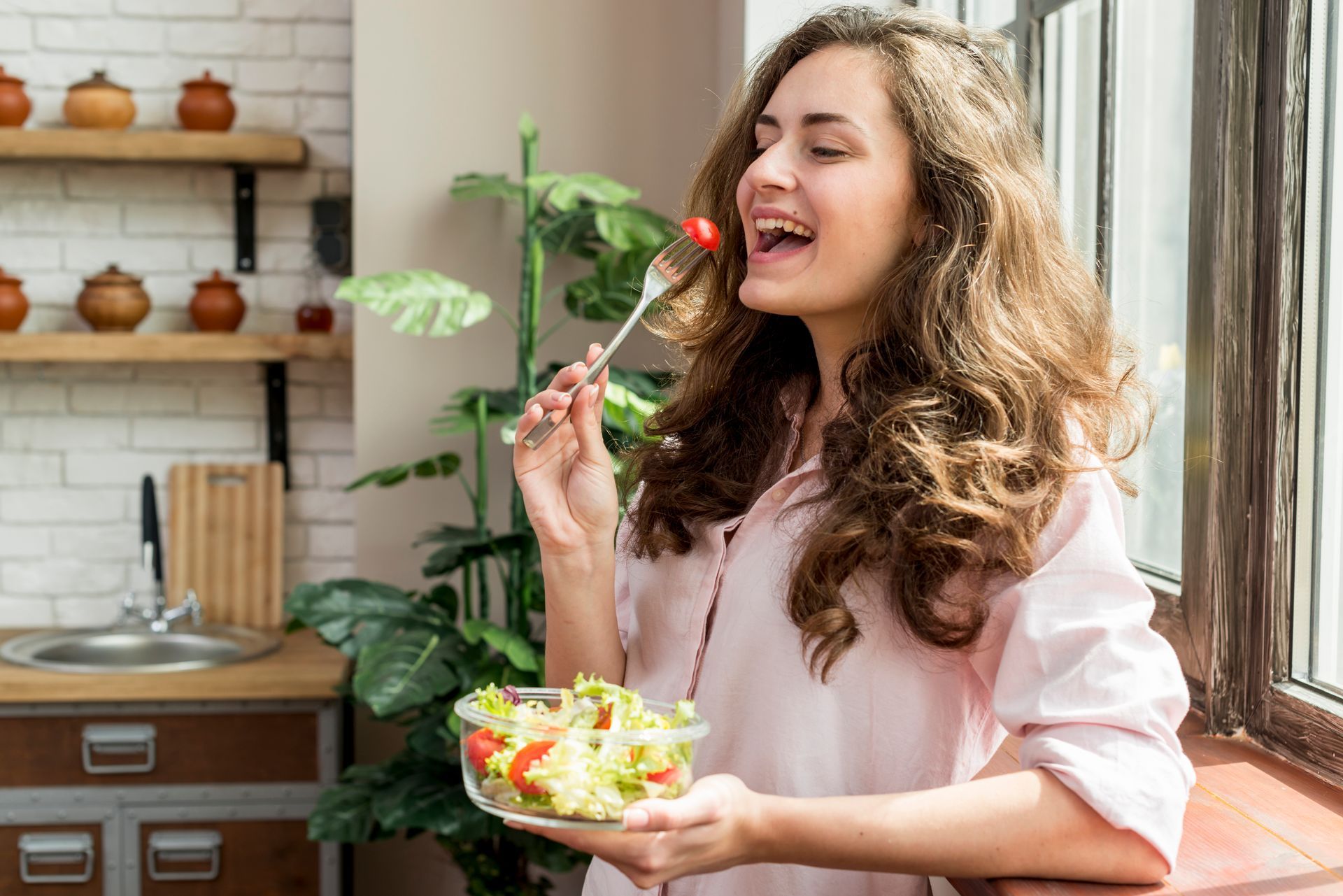 Woman smiling while eating a strawberry beside a bowl of salad in a bright kitchen