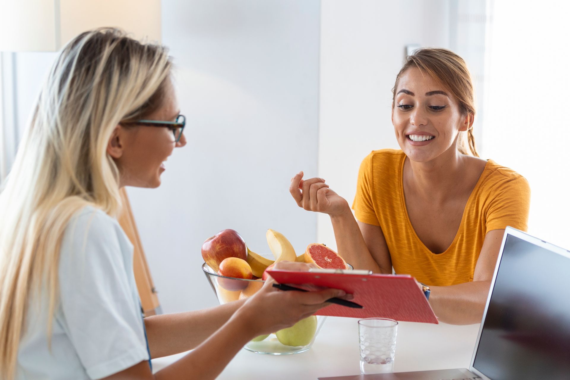 Two women talking at a table with fruit and a laptop, one holding a red clipboard in a bright office setting