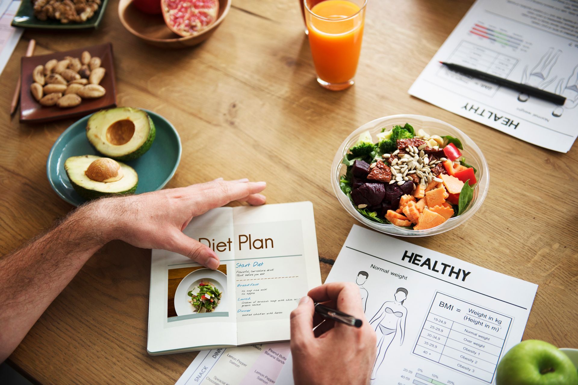 Hands planning a healthy meal plan with salad, avocado toast, fruit, and orange juice on a wooden table