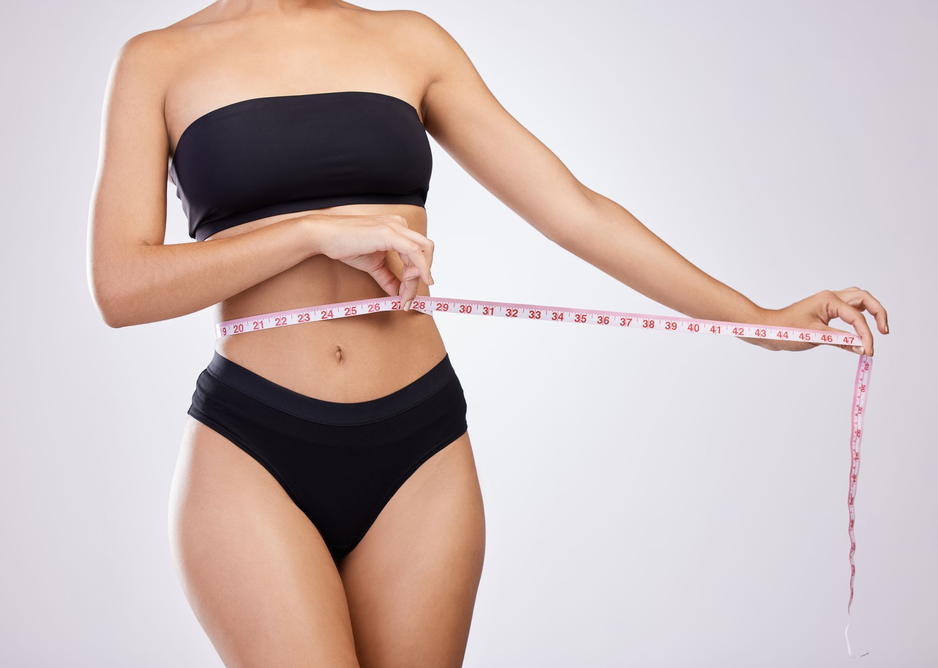 Person in black underwear measuring waist with a pink tape measure against a white background