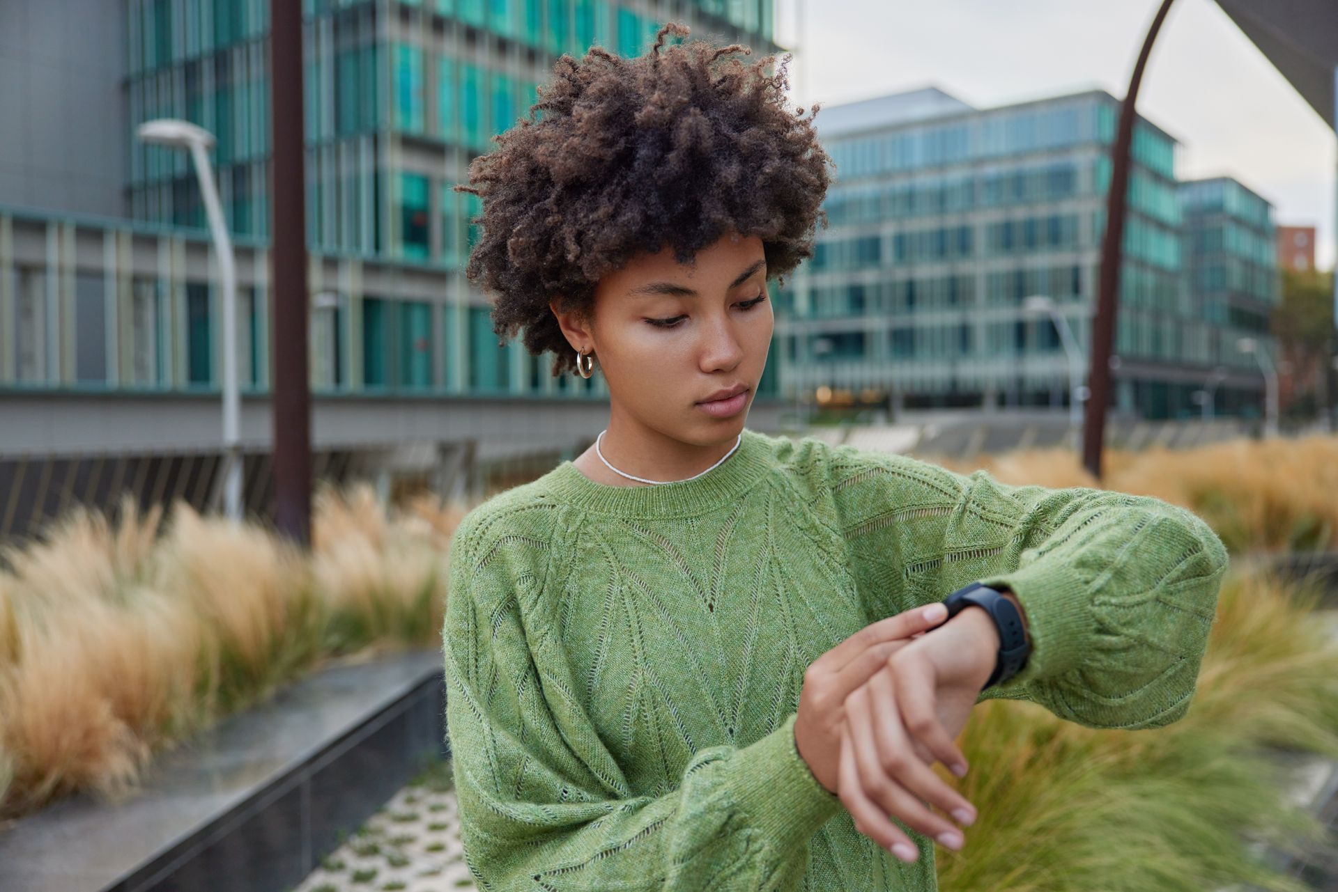 Person in a green sweater checking a smartwatch outdoors near modern buildings and grasses
