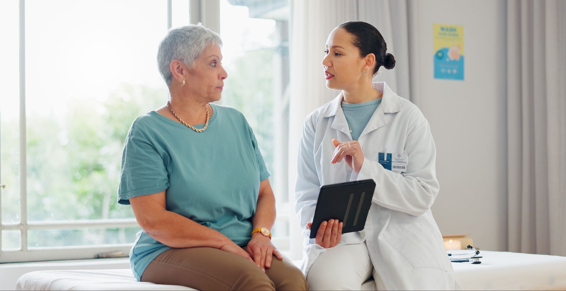 Doctor talking with patient in a bright clinic room, holding a tablet beside the exam table