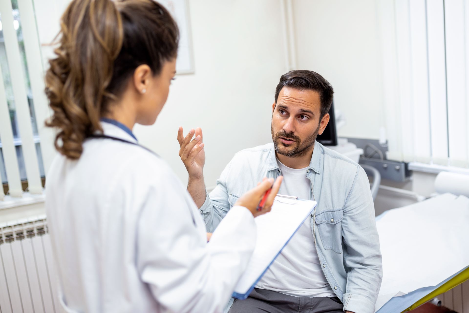 Doctor speaking with seated patient in a bright clinic room, holding a clipboard.