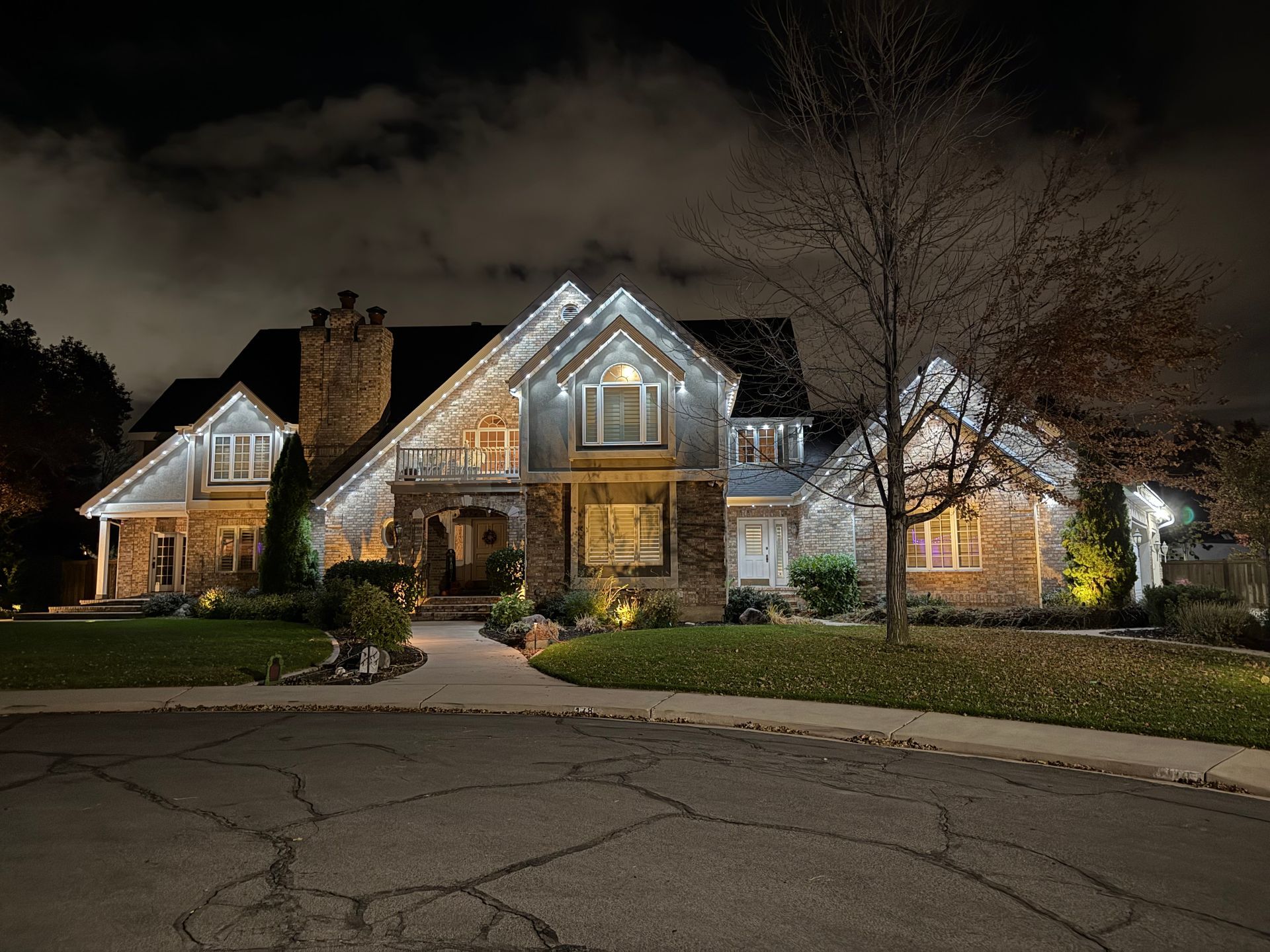 Large stone house with white holiday lights outlining the roof and windows, set against a night sky. Trees and a well-manicured lawn surround the home.