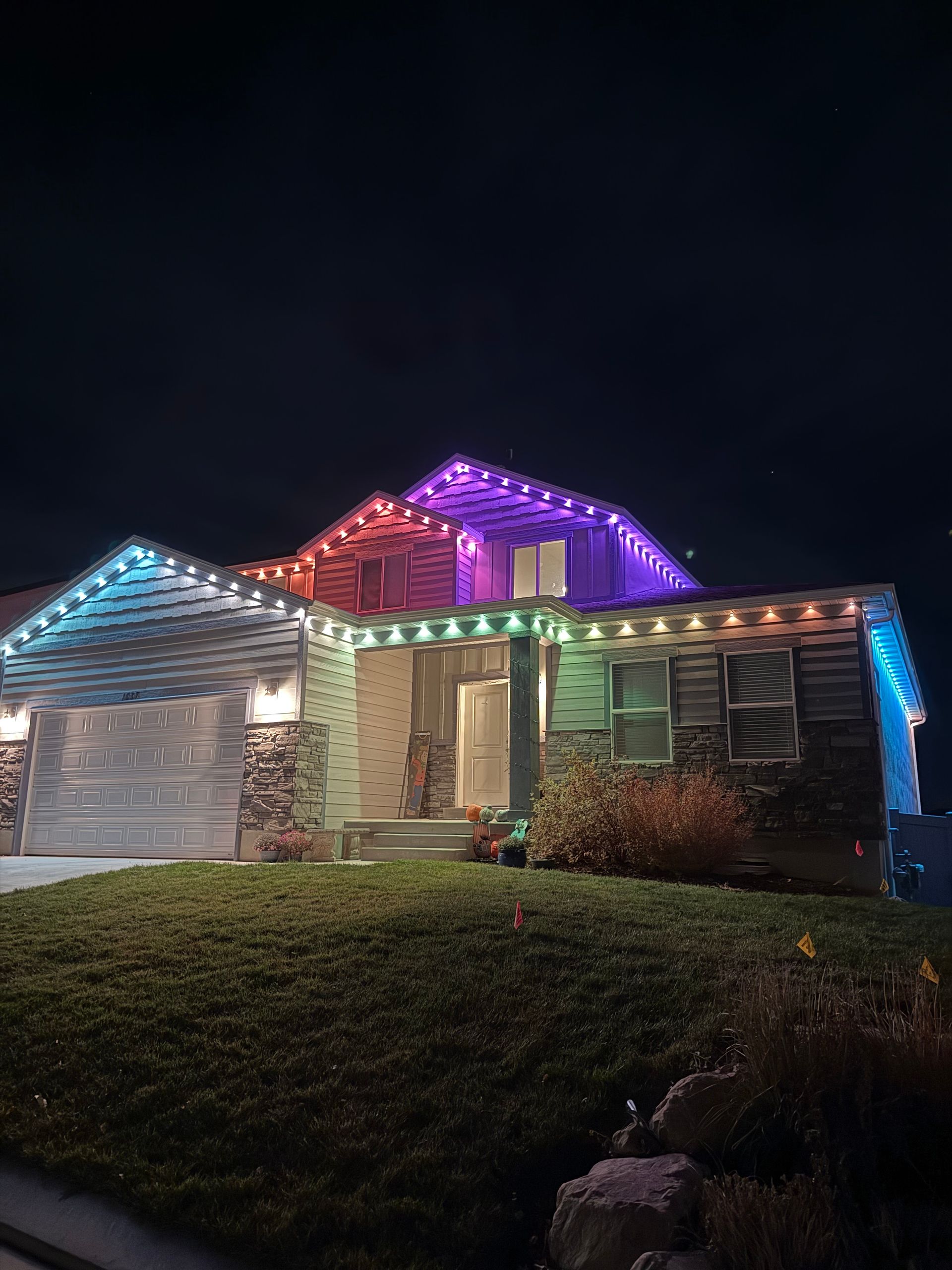 A suburban house illuminated by colorful, festive LED lights outlining the roof edges against a dark night sky, creating a vibrant, cheerful atmosphere.