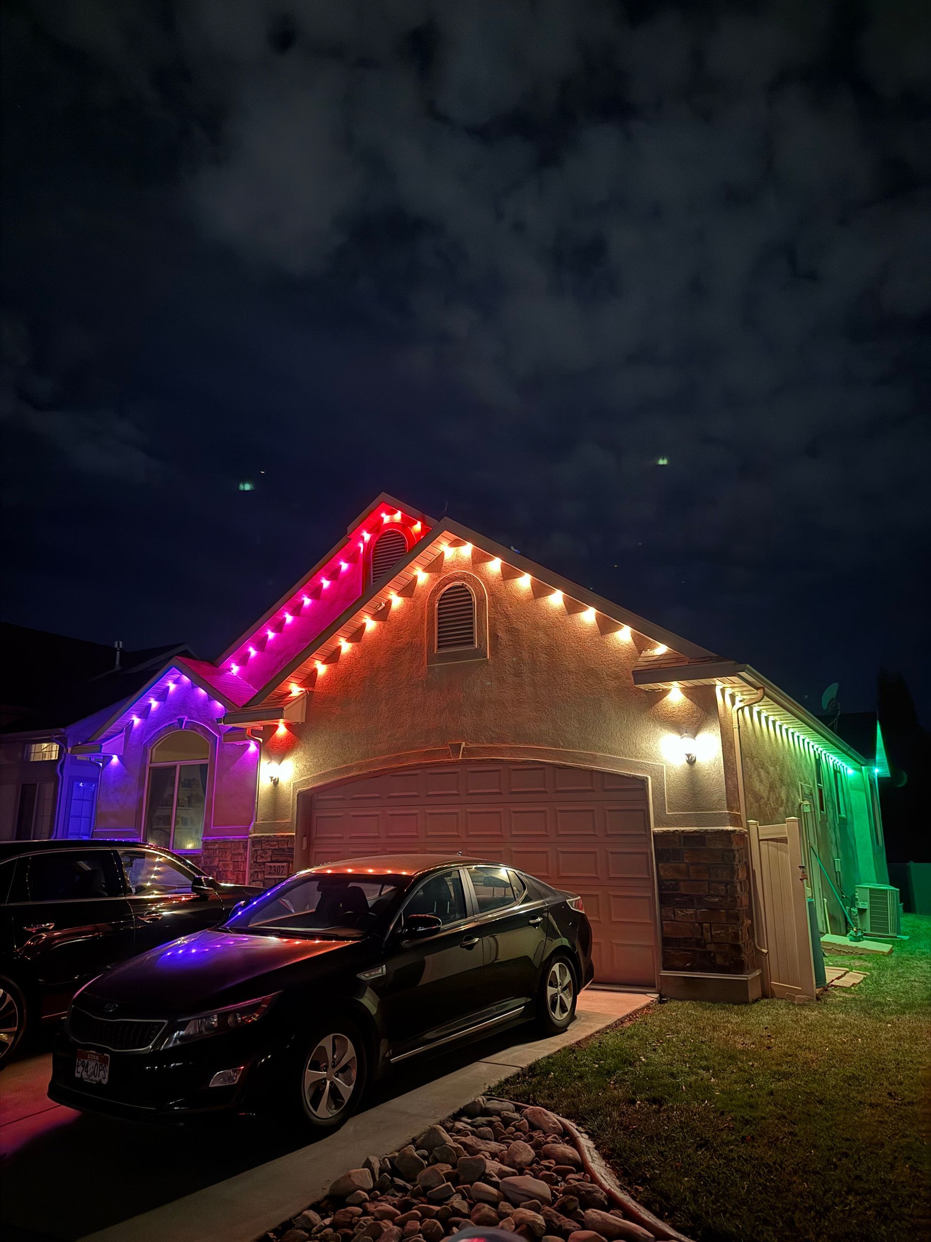 A house lit with colorful LED lights on the eaves, transitioning from purple to green. A glowing atmosphere contrasts with the night sky.