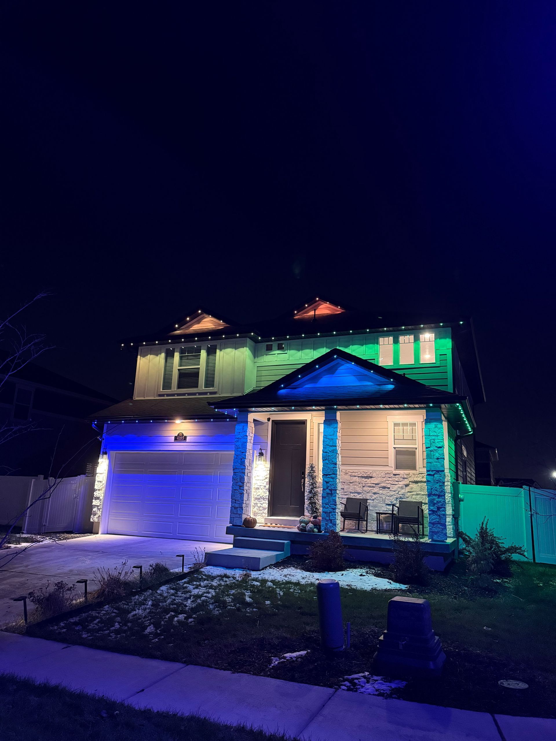 Two-story house at night illuminated with blue and green lights, creating a festive, vibrant atmosphere. The siding and garage are visible with a clear, dark sky.