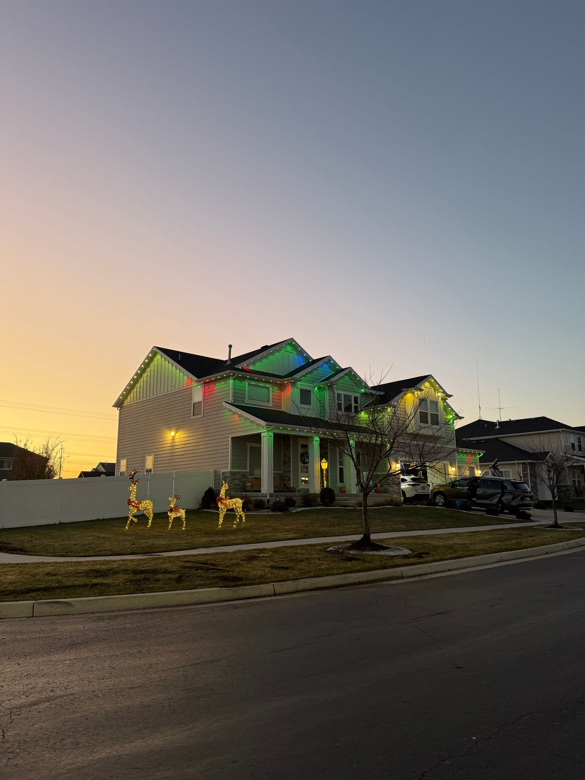 A house adorned with festive multicolored Christmas lights at sunset. Three lit reindeer decorations stand on the lawn, creating a cheerful holiday scene.