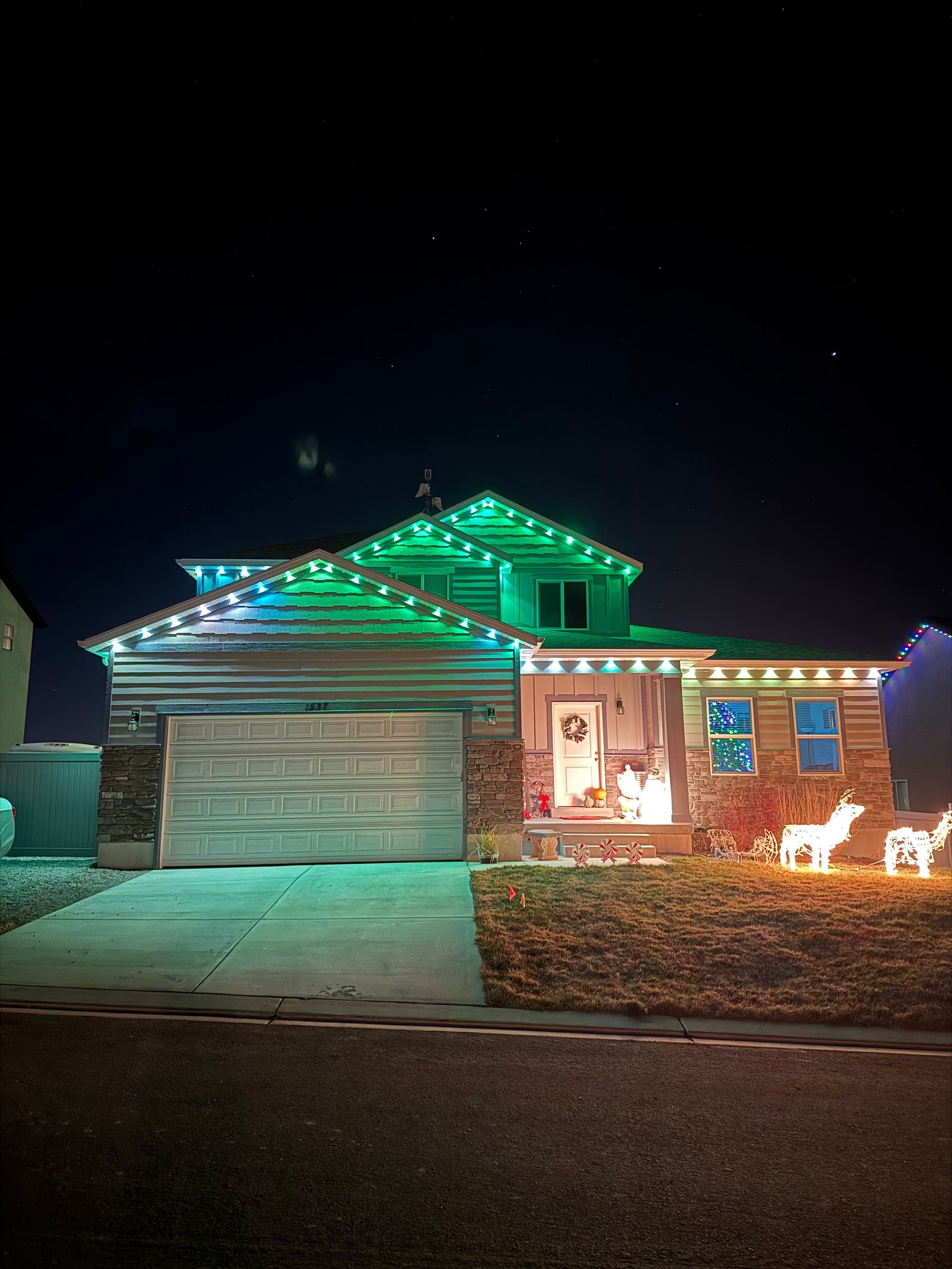 A festive suburban house at night, adorned with green and white Christmas lights on the roof. Glowing reindeer decorations adorn the front yard.
