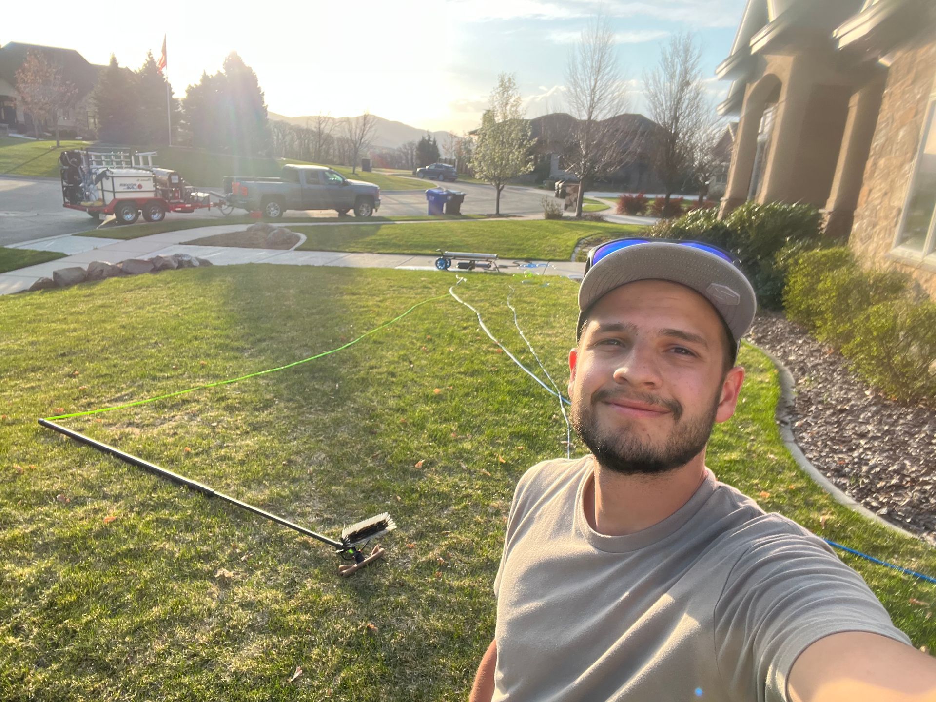 A man in a gray cap smiles for a selfie in a sunlit yard. A hose and equipment are on the grass. A truck and trailer are in the driveway.