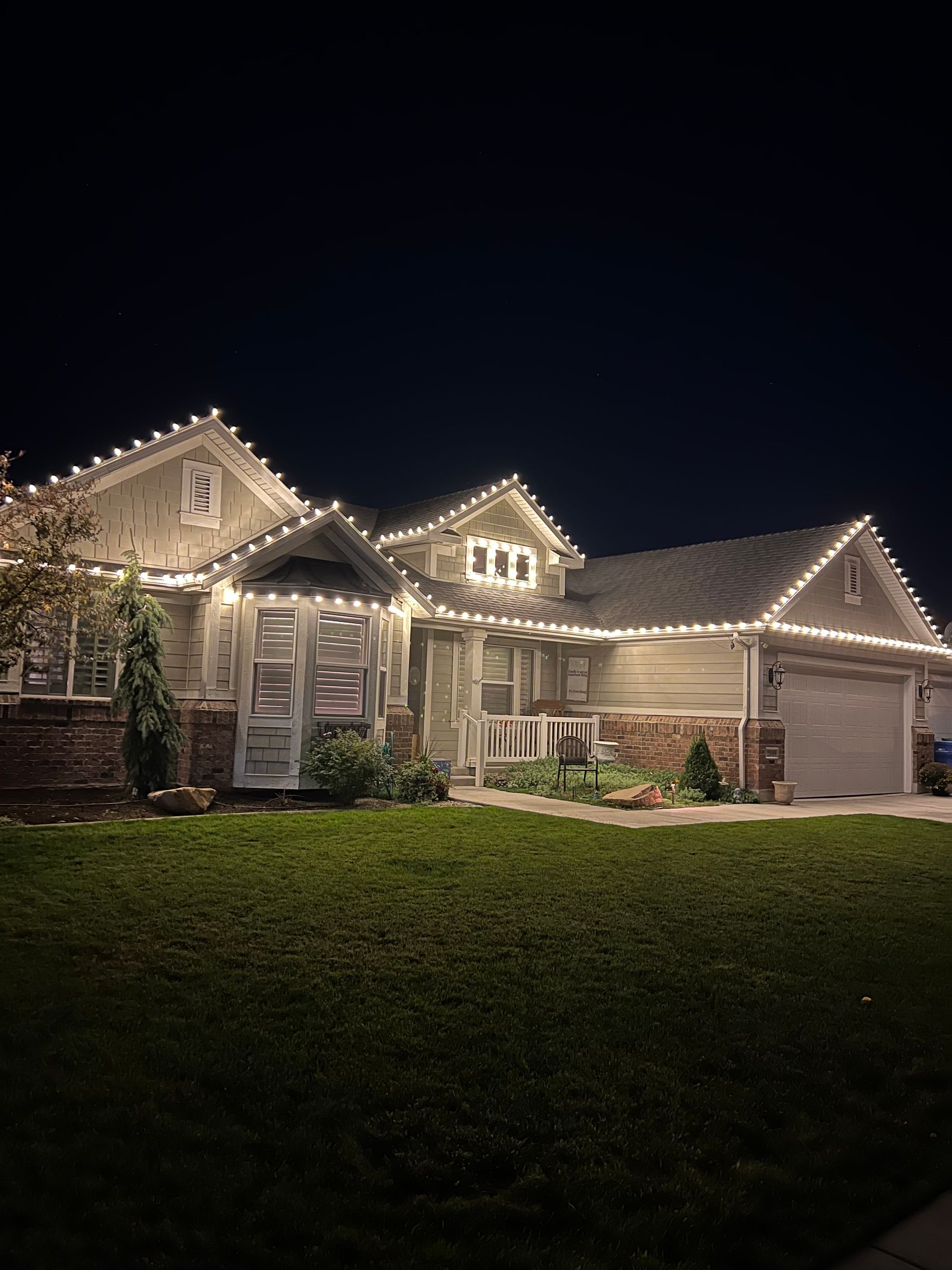 A cozy house at night adorned with warm white string lights along the roofline, illuminating the yard and creating a festive and welcoming atmosphere.