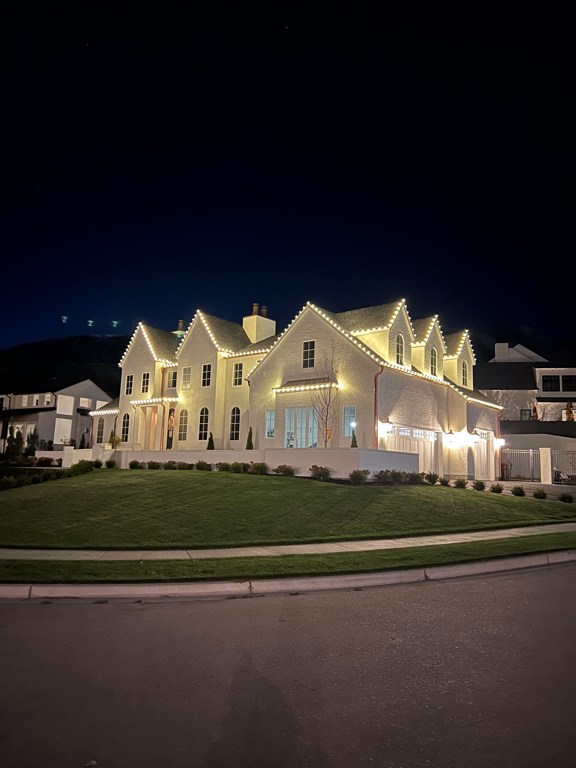 A large house adorned with glowing string lights stands illuminated against the night sky. The warm lights highlight the architecture, creating a festive ambiance.