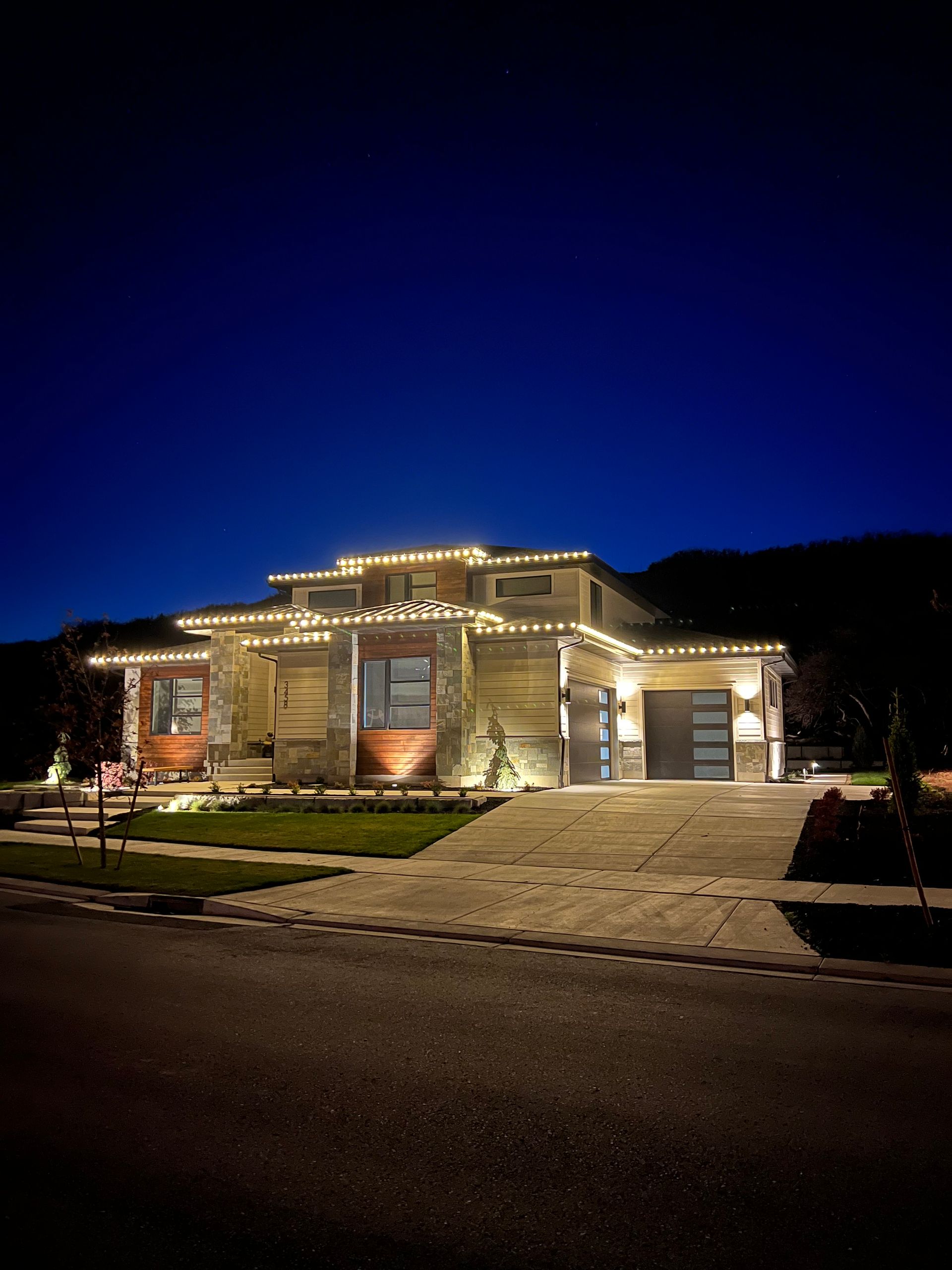A modern house at night, outlined with festive white string lights, features a stone facade and large garage doors, set against a dark blue sky.