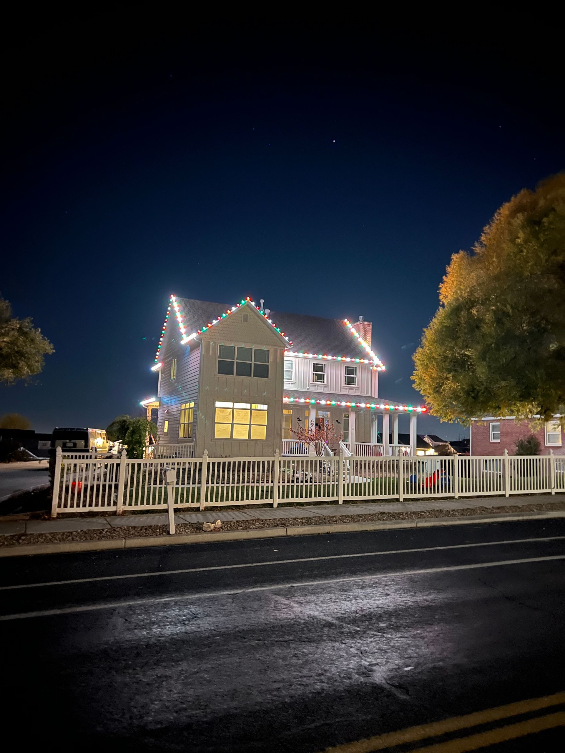 A cozy two-story house adorned with colorful holiday lights along the roof and porch. A bright night sky and large trees enhance the festive atmosphere.