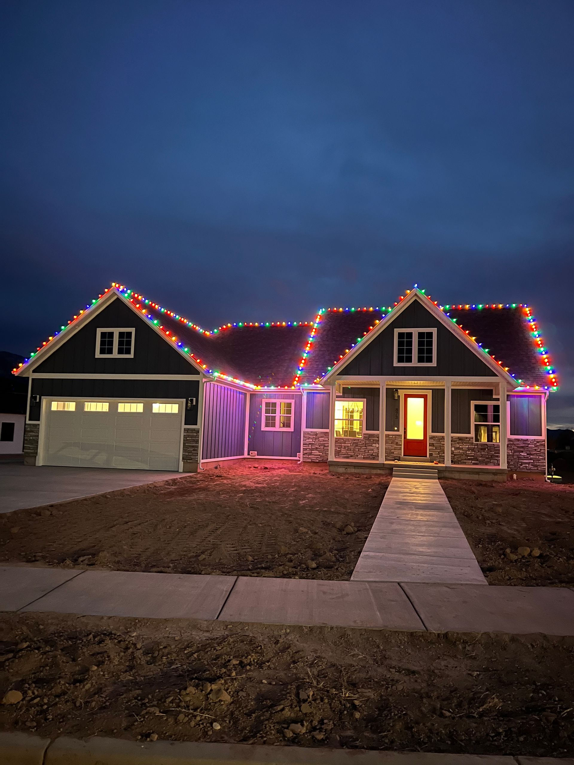 A cozy house is adorned with colorful Christmas lights outlining its roofline. The sky is dark, and warm indoor light glows from the windows, creating a festive atmosphere.