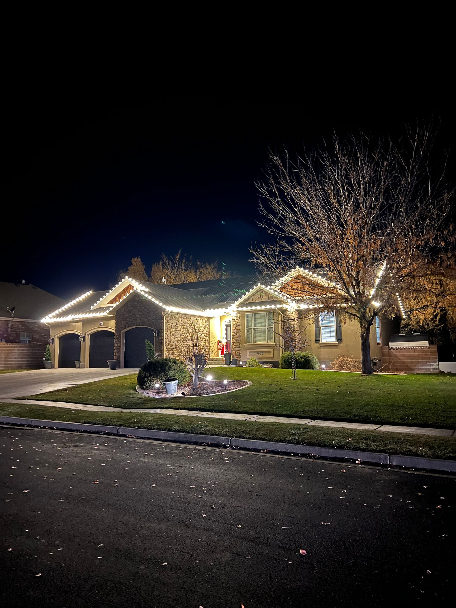 Single-story house at night with warm stone facade and Christmas lights outlining the roof and tree. Festive and welcoming atmosphere.