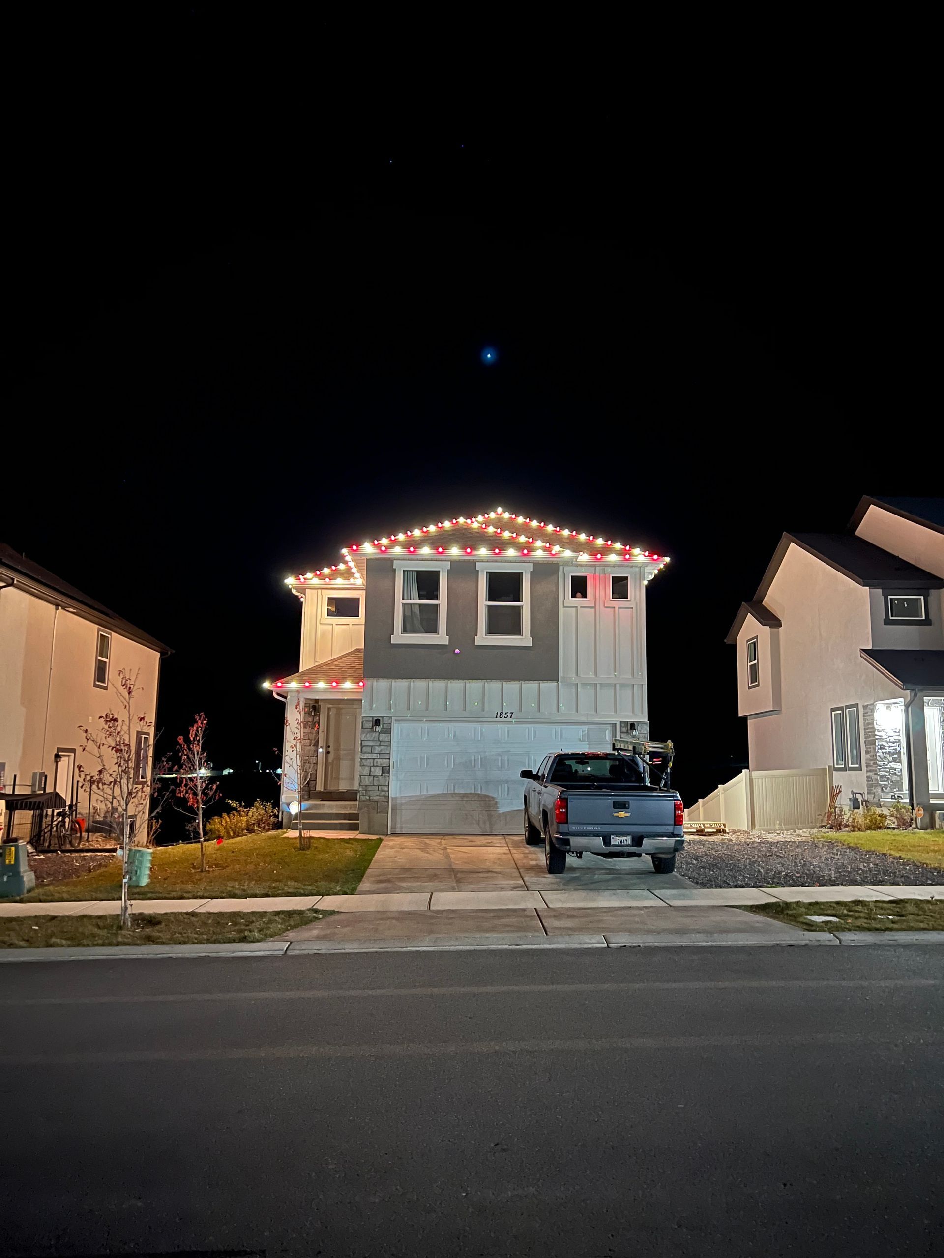 A two-story house with colorful Christmas lights on the roof is flanked by similar homes at night. A truck is parked in the driveway, with a dark, starry sky above.
