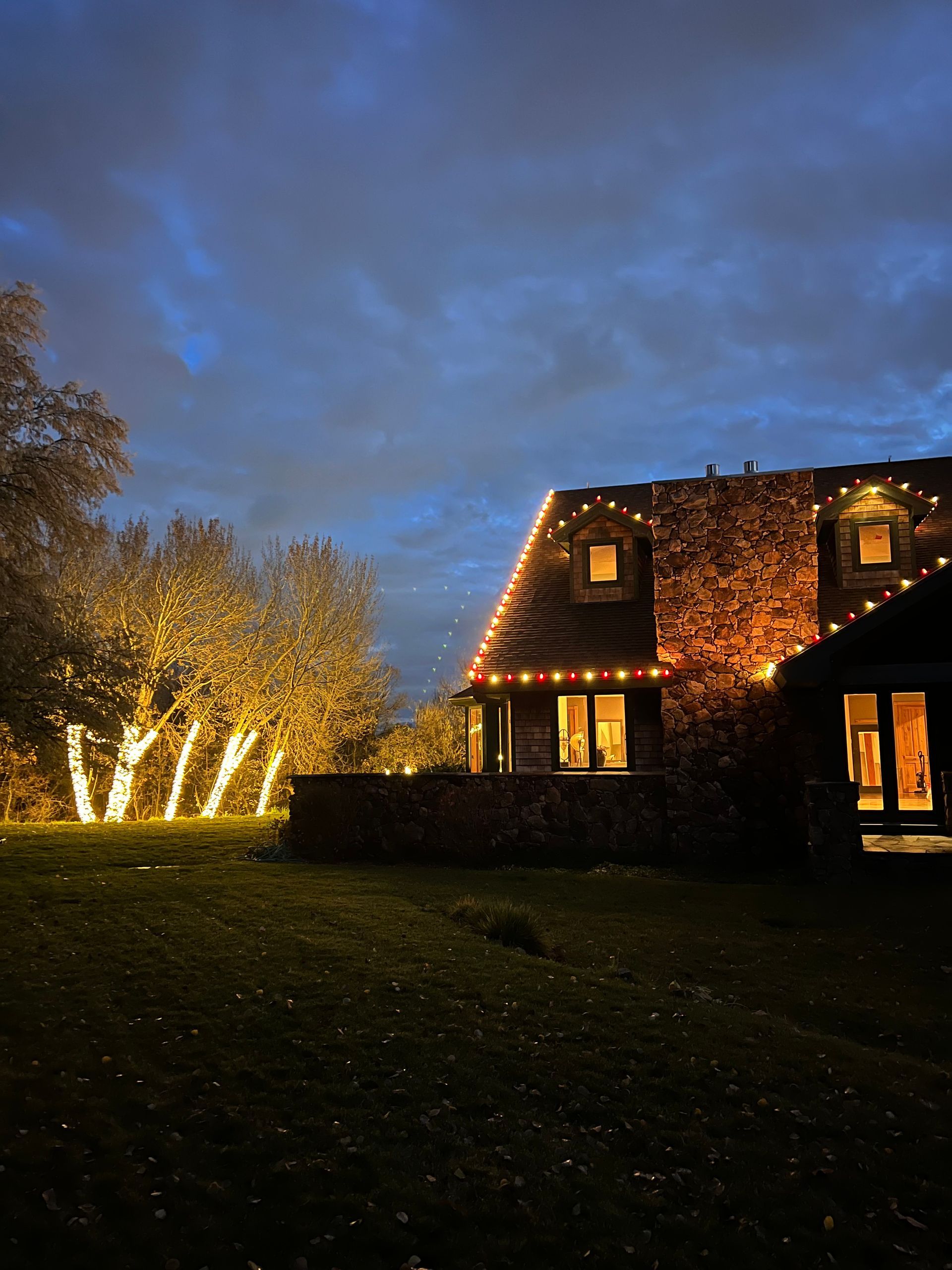 Cozy house adorned with festive string lights along the roof at dusk. Illuminated trees stand nearby, creating a warm, inviting glow against the darkening sky.