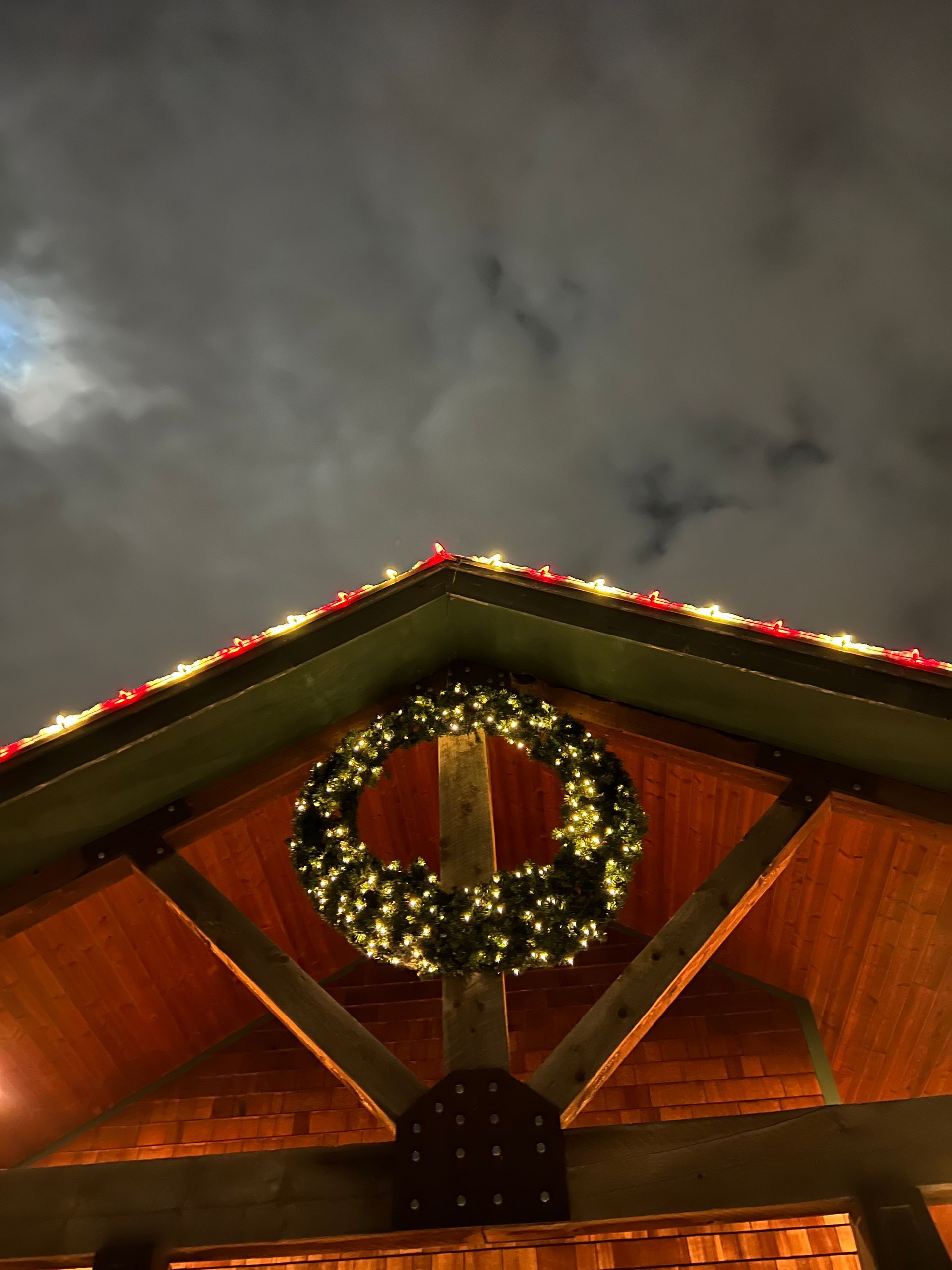 Wooden structure with a large, illuminated green wreath hanging under its peak. Rooflines adorned with red and white lights against a cloudy night sky.
