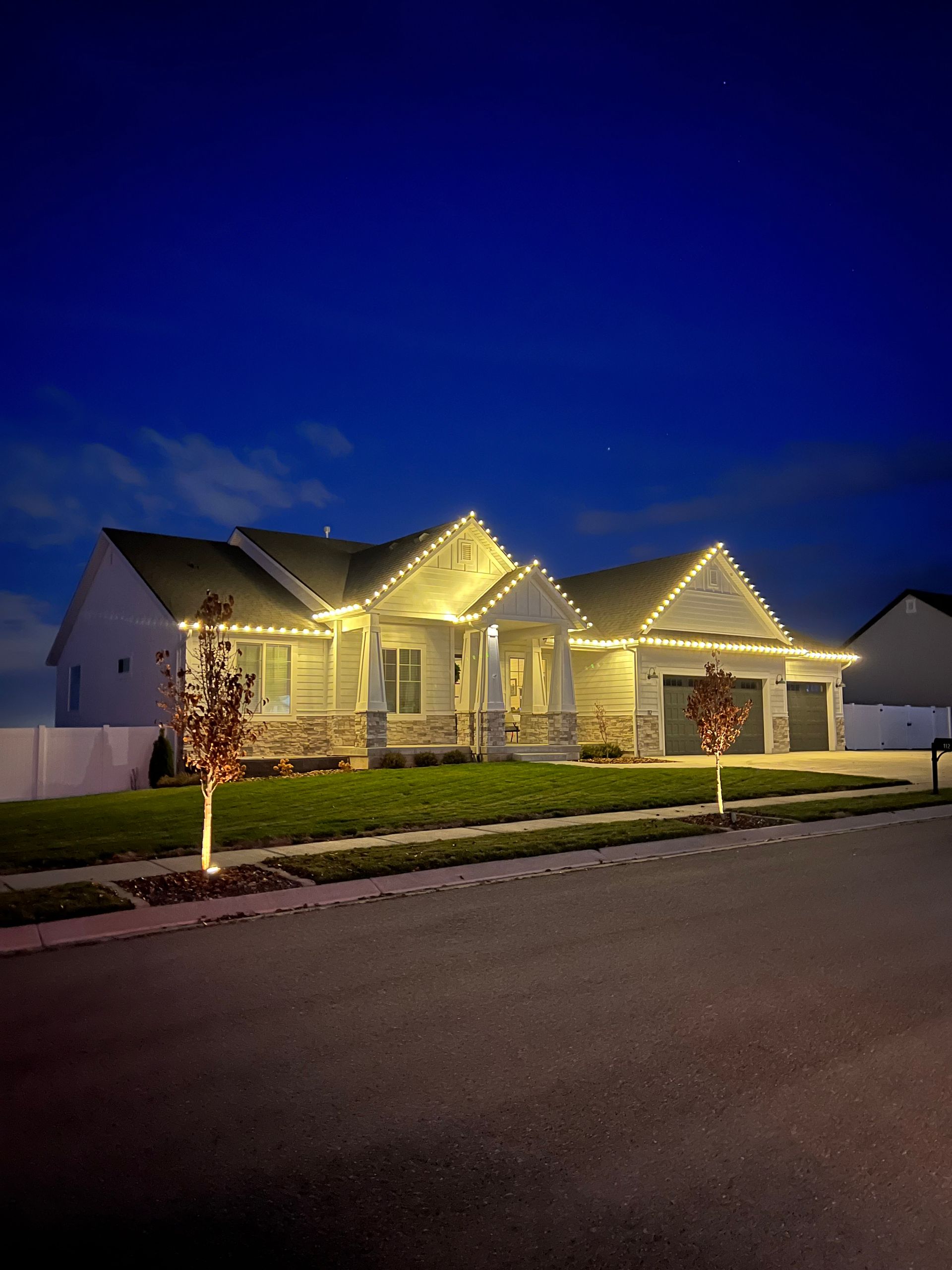Single-story house at dusk, adorned with warm white Christmas lights outlining the roof. Neatly trimmed lawn, flanked by young trees also wrapped in lights.