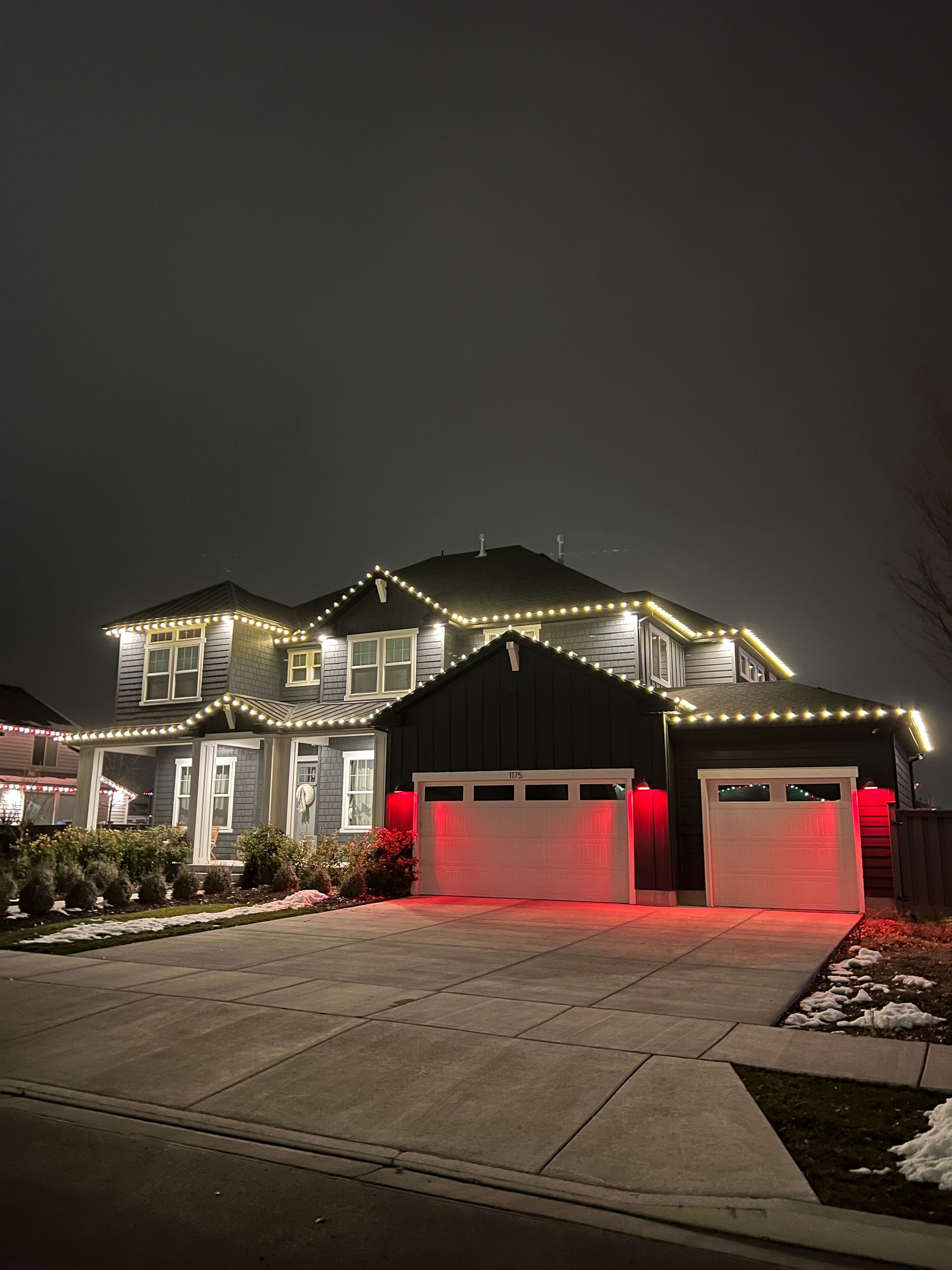 Two-story house at night, adorned with white string lights and soft red lighting near the garage. A festive, warm, and inviting atmosphere.